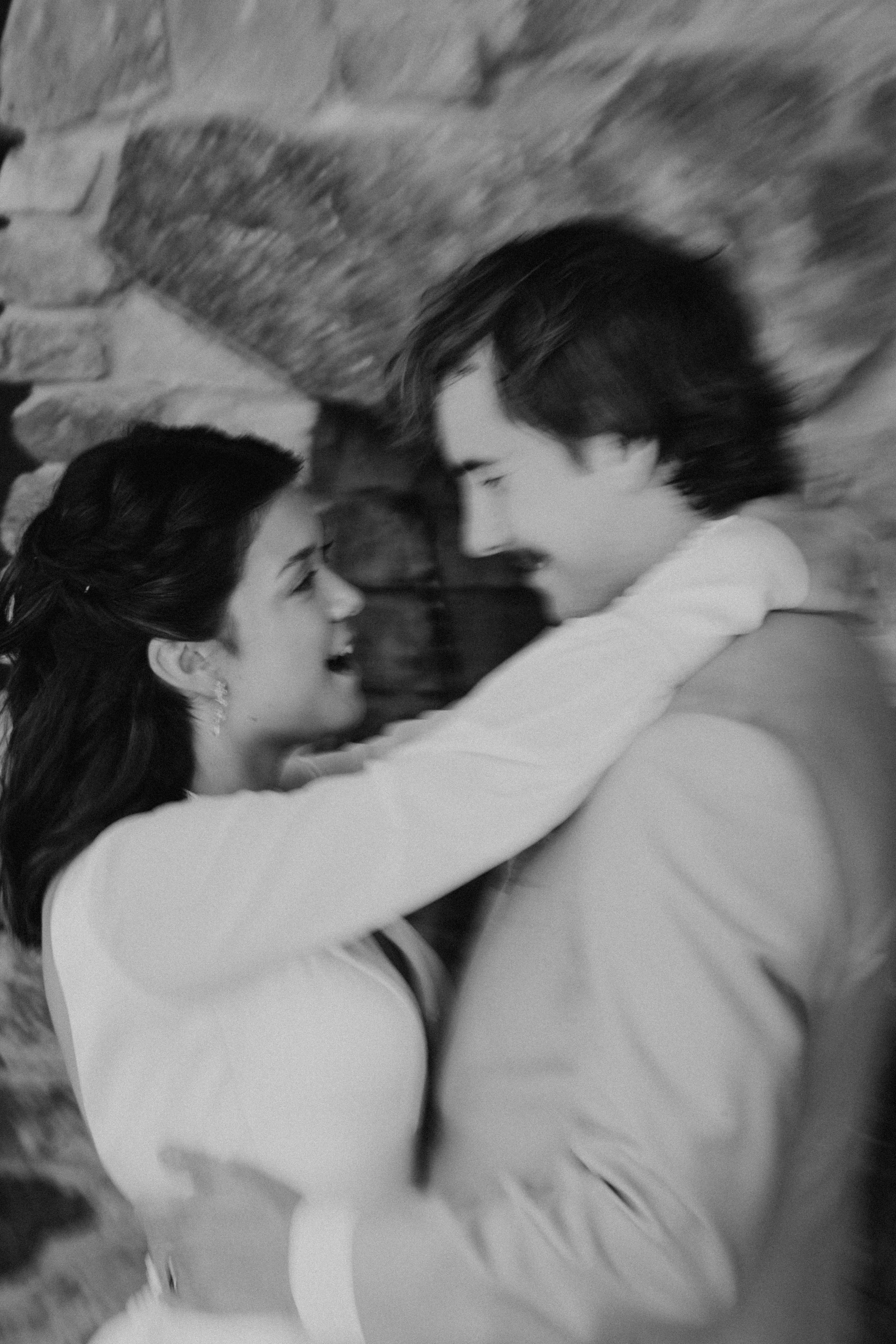 A black-and-white photo of a man and woman embracing each other closely, looking into each other's eyes with a stone wall in the background.