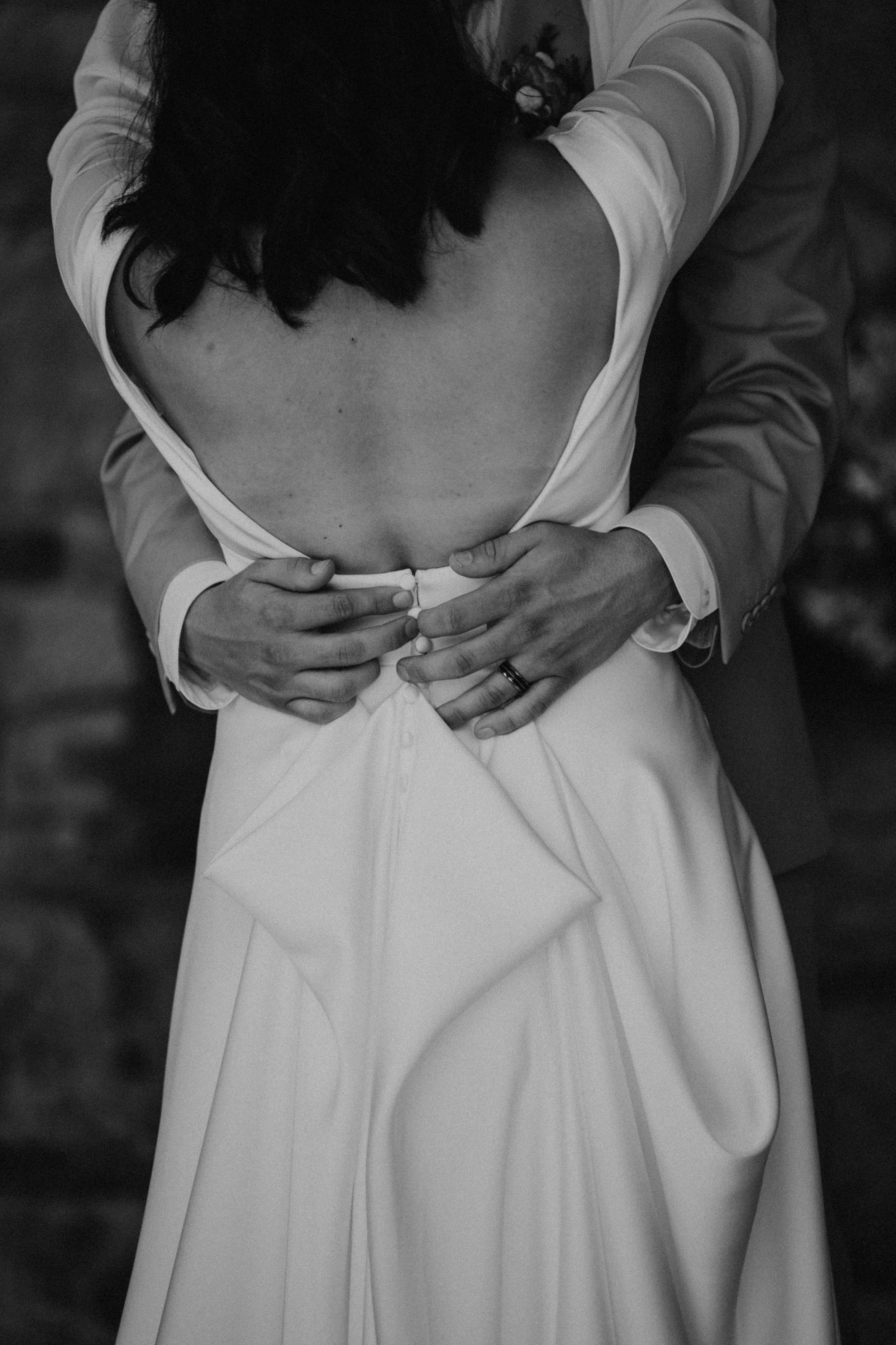 A black and white photo showing a man and woman embracing, with the man's arms around the woman's waist. The woman is wearing a wedding dress and the man is in a suit. Their faces are not visible.