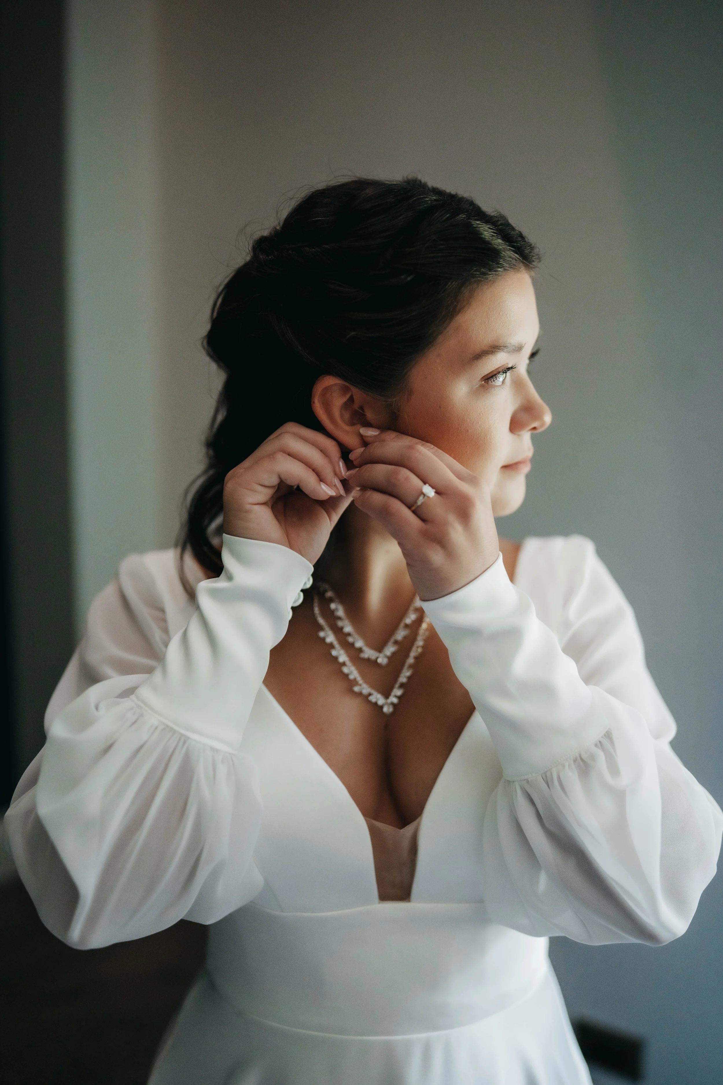 A bride with dark hair in a white dress is putting on earrings, wearing a diamond ring and a layered pearl necklace, looking out the window.