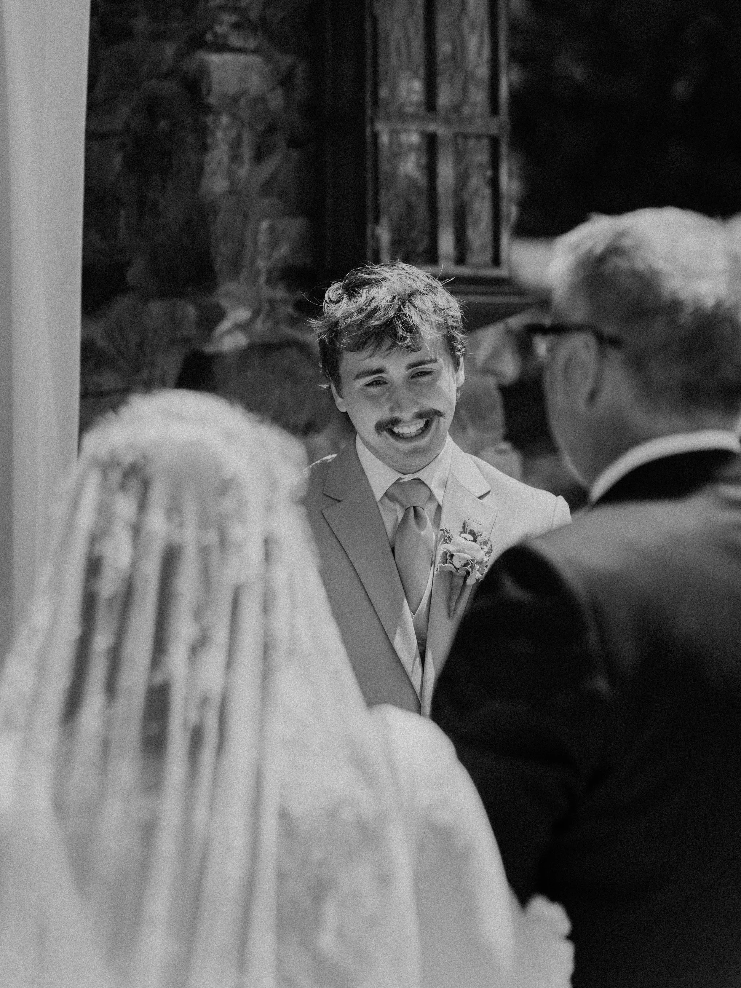 Black and white photo of a young groom with a mustache and wavy hair, smiling inside a church, facing an older man in glasses, and with a woman in a veil nearby.