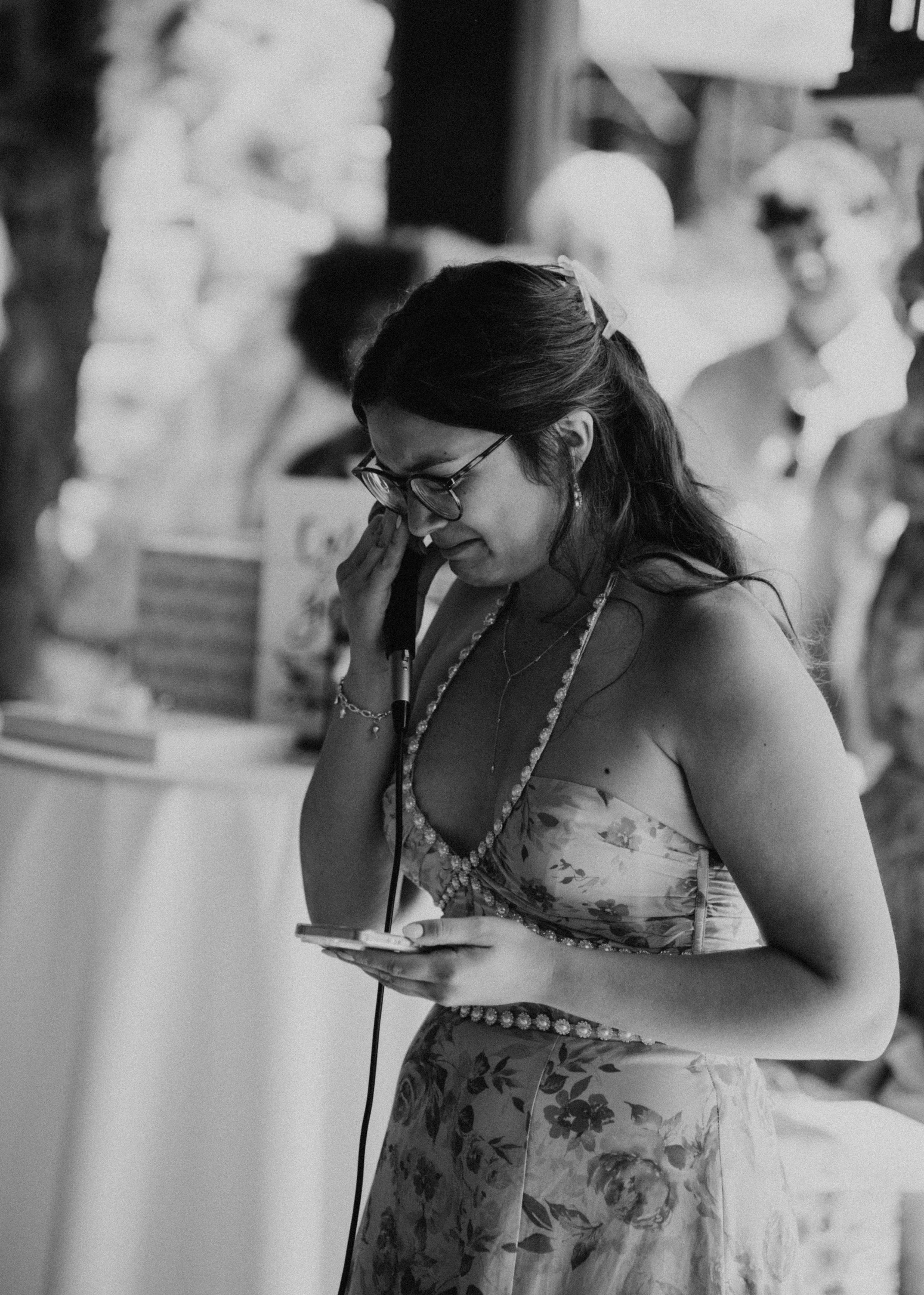 A woman in a floral dress, wearing glasses and jewelry, is talking on the phone while looking at her phone, at what appears to be a social gathering or celebration.