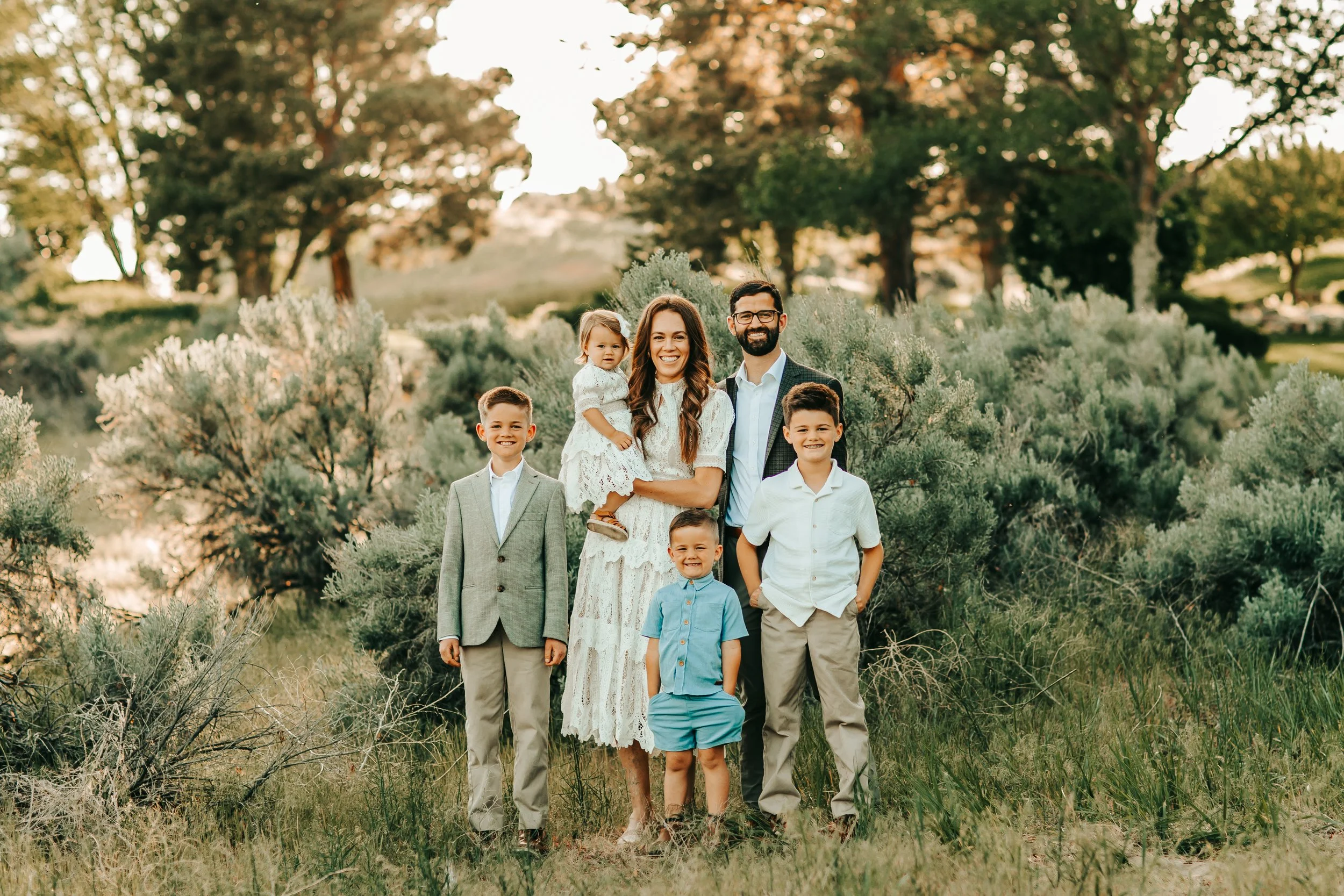 Family photo outdoors with six people, two adults and four children, standing among greenery and trees during sunset.