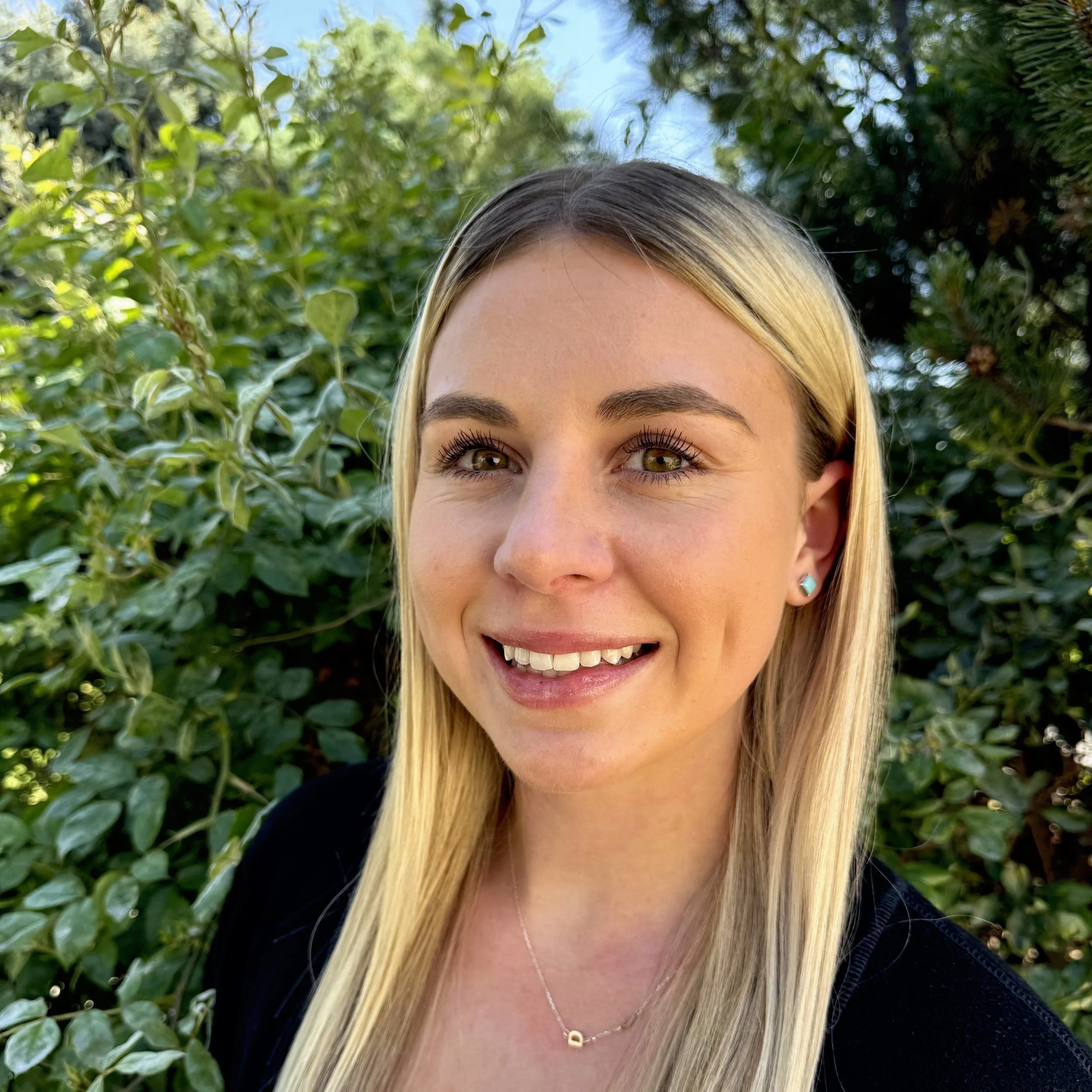 A young woman with long blonde hair, light makeup, and wearing a black top and turquoise earrings, smiling outdoors with green foliage and blue sky in the background.