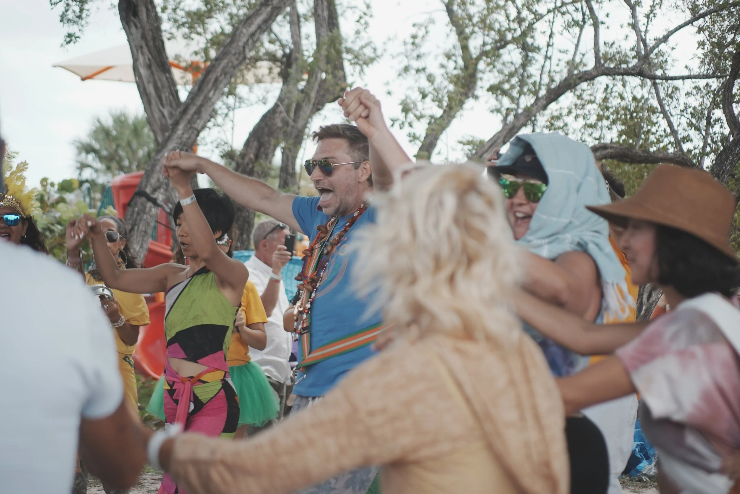 People dancing and celebrating outdoors, wearing colorful clothing and accessories, with trees and a slide in the background.