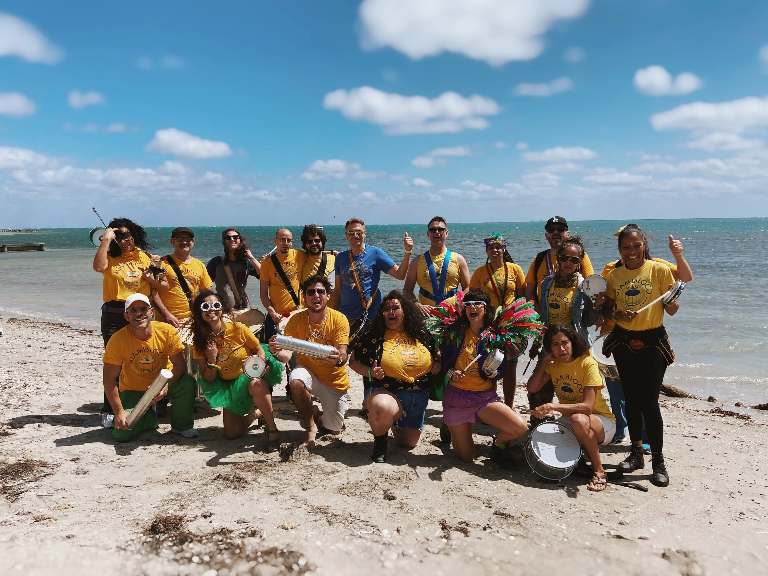 A group of people on a beach wearing yellow t-shirts with a logo, holding musical instruments, and posing for a photo under a partly cloudy sky.