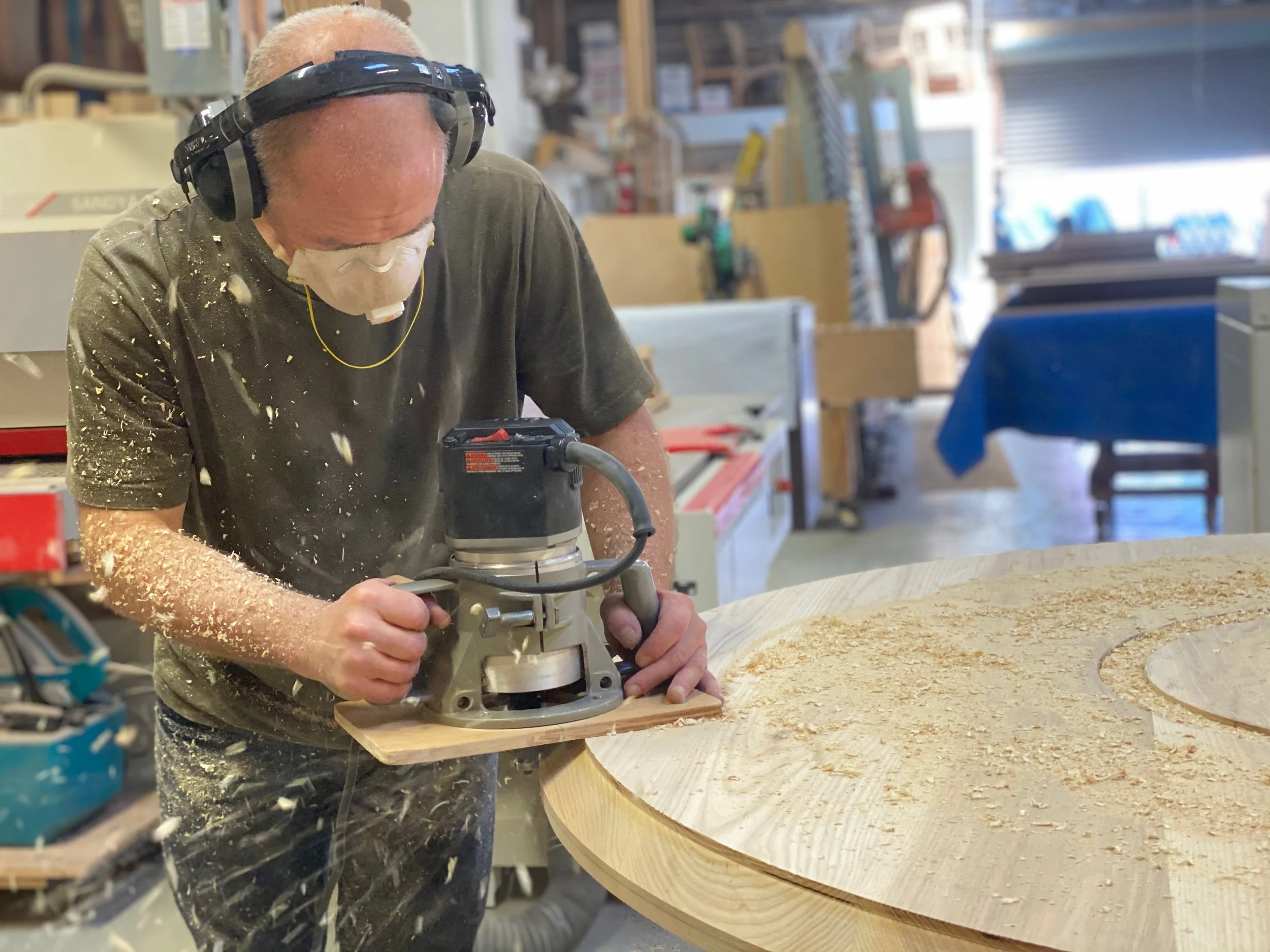 A man using a wood router on a large circular wooden piece in a woodworking shop, wearing ear protection and a face mask, with sawdust flying around.