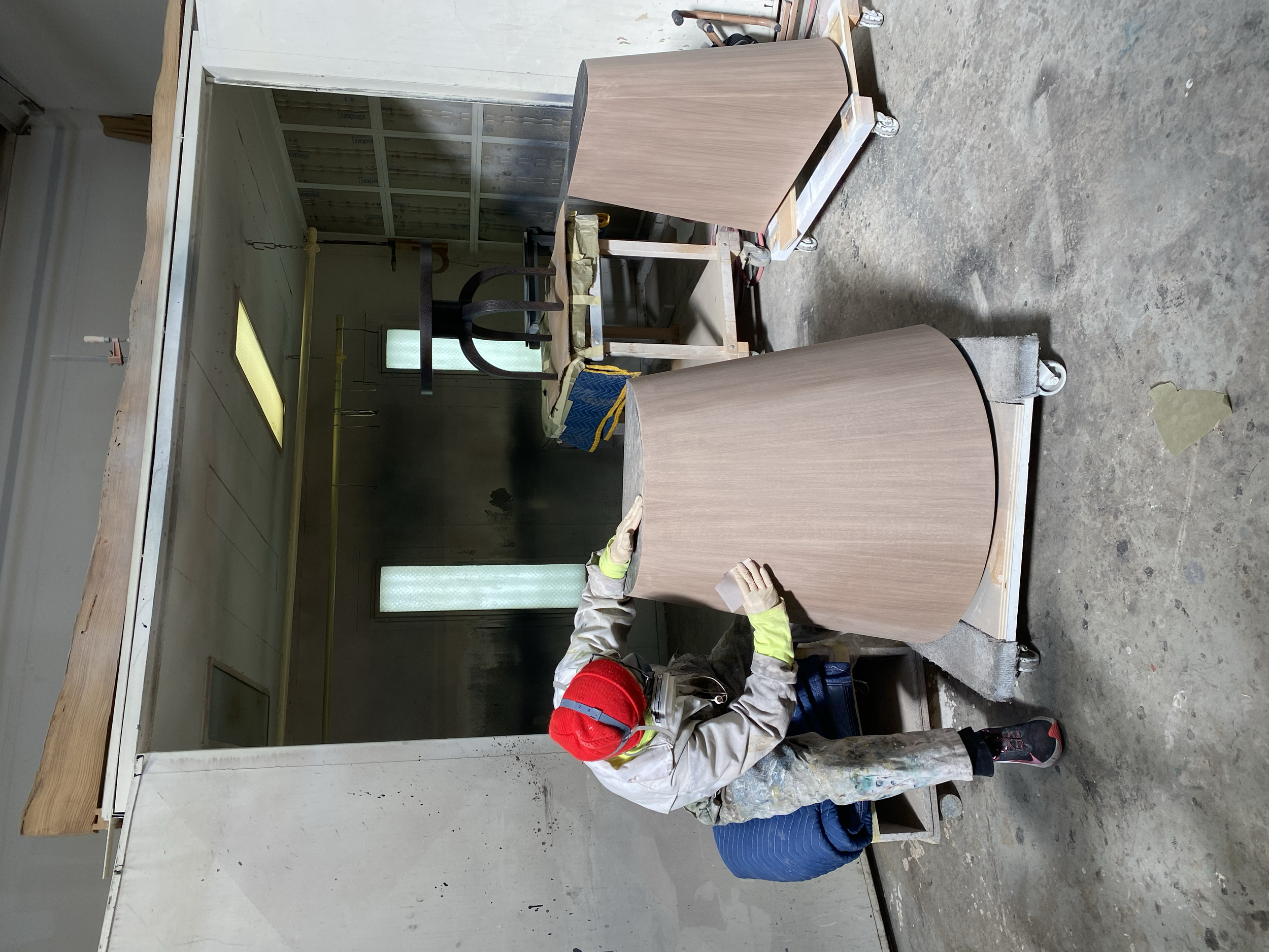 A worker in a workshop sanding a piece of curved wooden furniture, wearing protective gloves, a red beanie, and paint-splattered clothes, with woodworking tools and equipment in the background.