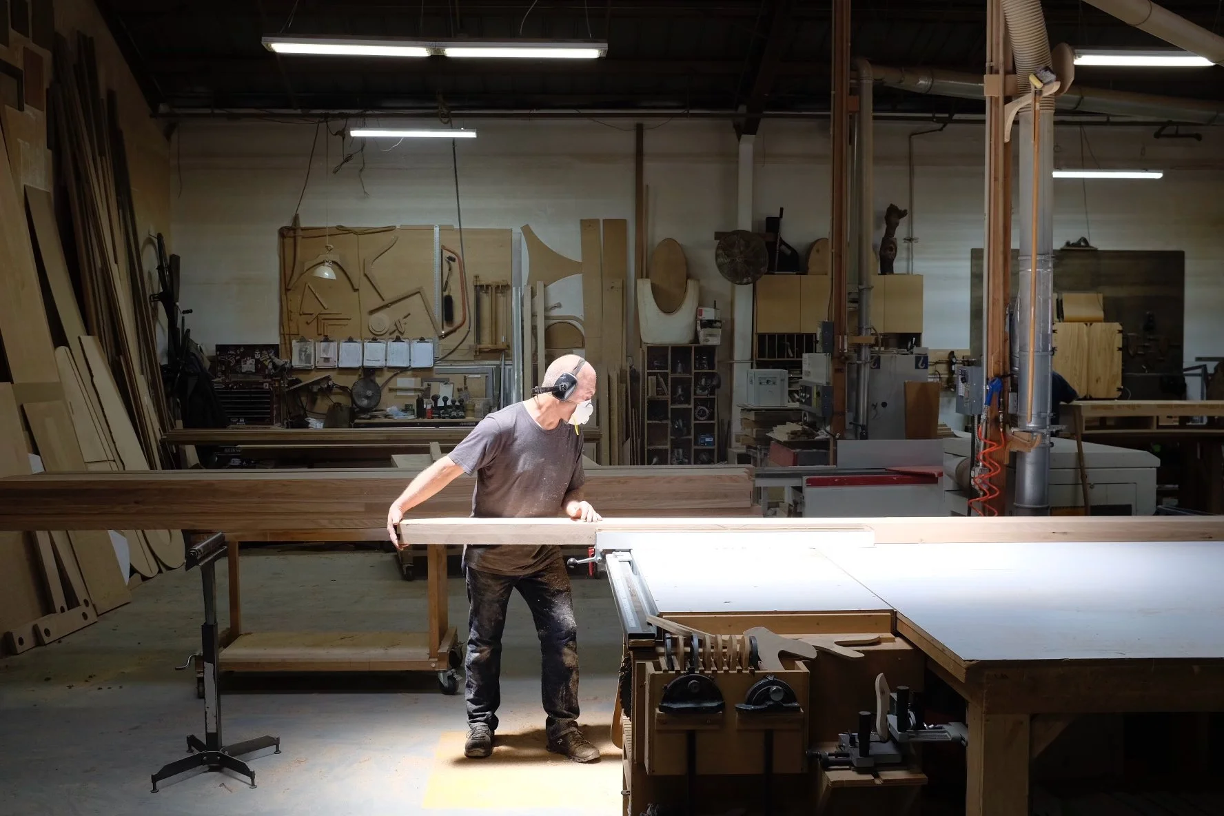 A person cutting wood in a woodworking shop, wearing ear protection and a face mask, surrounded by woodworking tools and equipment.