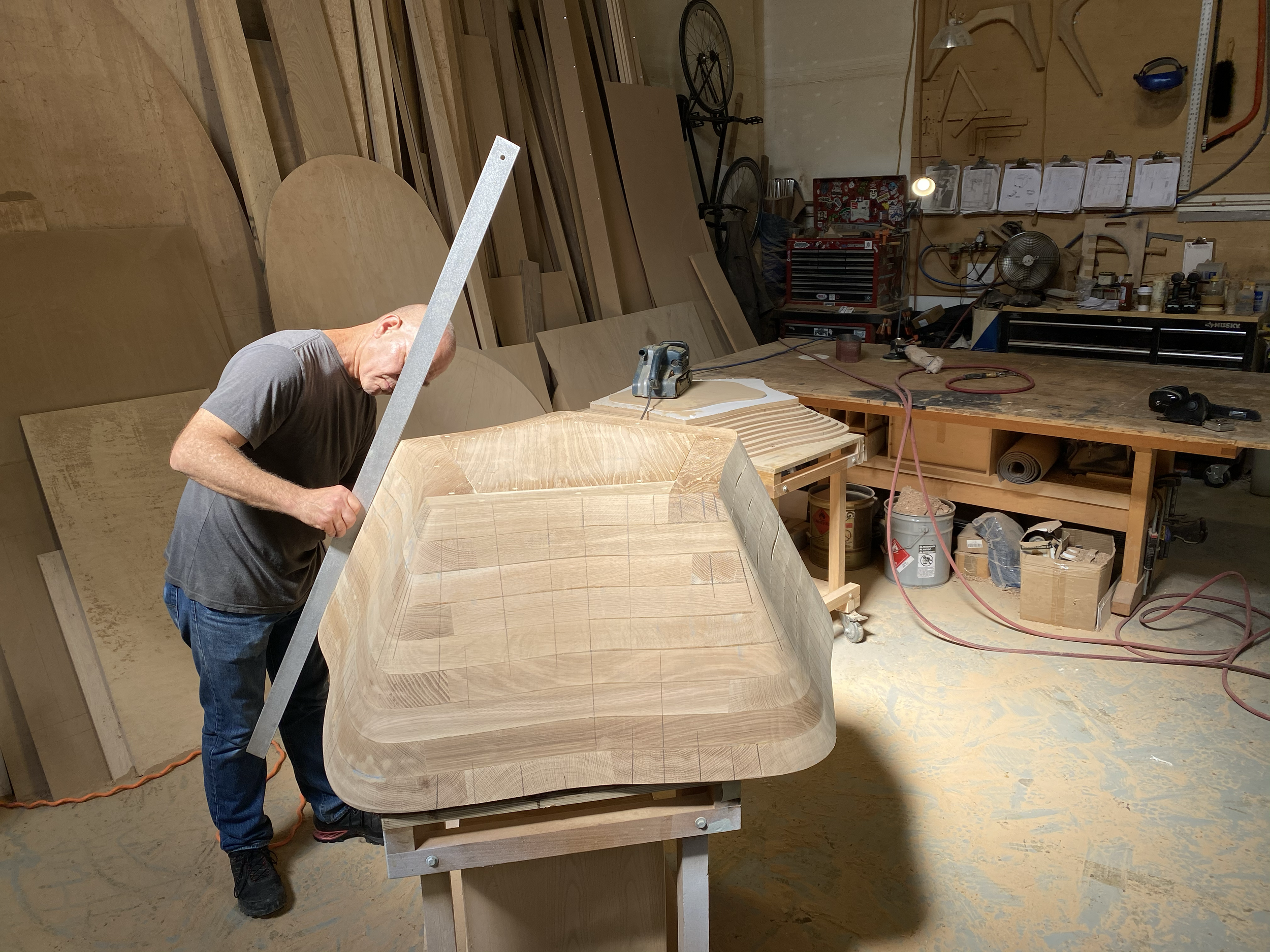 A man working in a woodworking shop, shaping a large piece of wood on a workbench, surrounded by woodworking tools and materials.