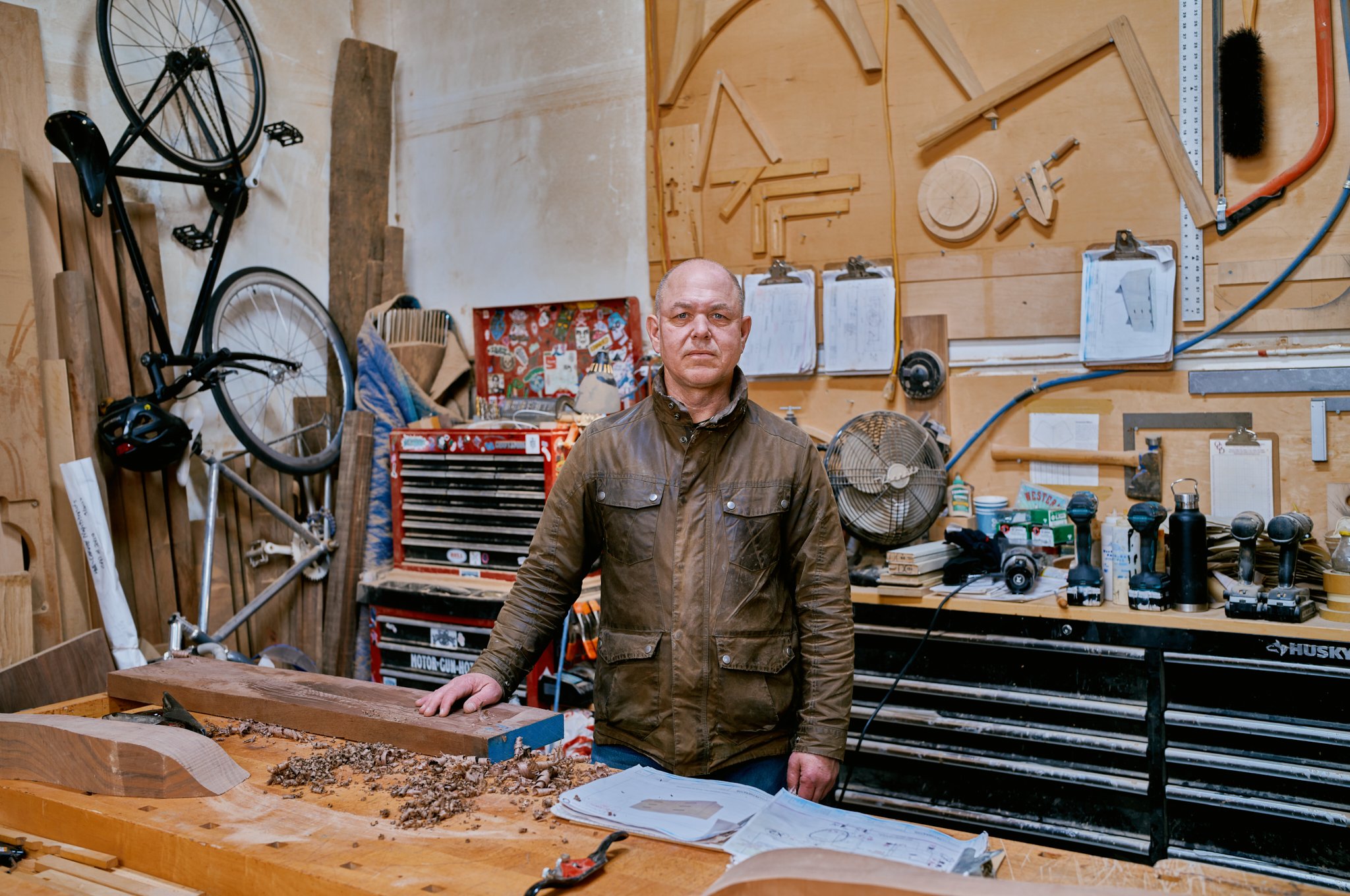 A man in a brown leather jacket standing in a woodworking workshop, with tools, wood pieces, and plans on the workbench and colorful workshop wall behind him.