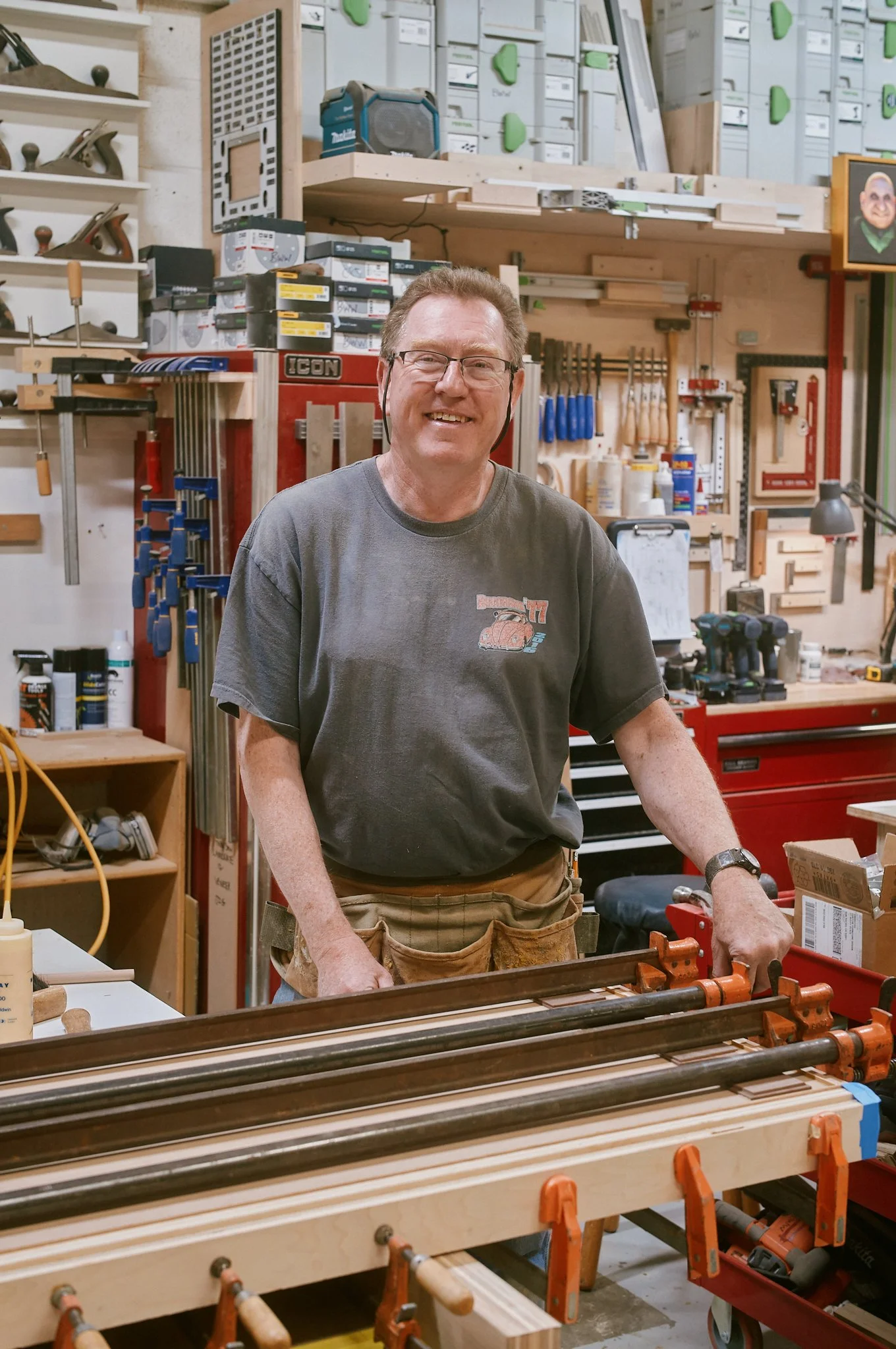 A man smiling in a woodworking workshop, surrounded by tools and lumber, holding a clamp.