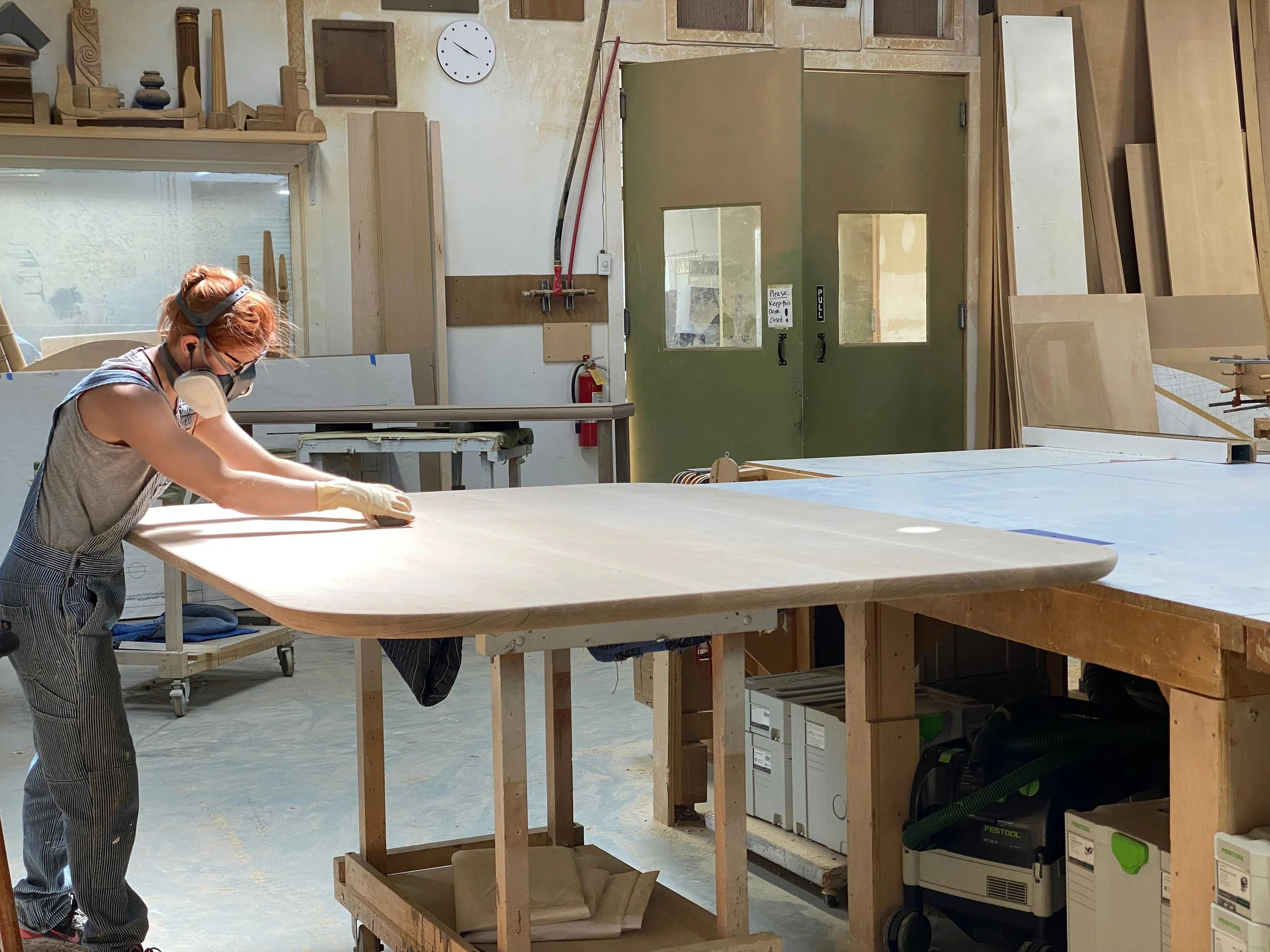A woman working on a woodworking project in a workshop, sanding a large wooden surface.
