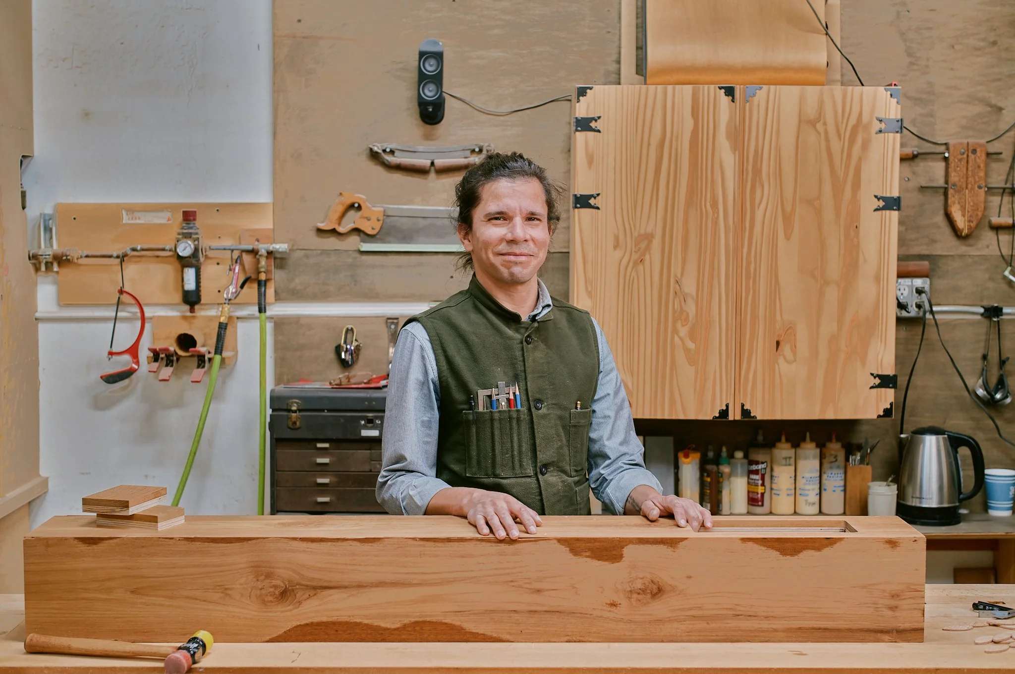 A man in a workshop standing behind a wooden workpiece, with woodworking tools and supplies on shelves and walls behind him.