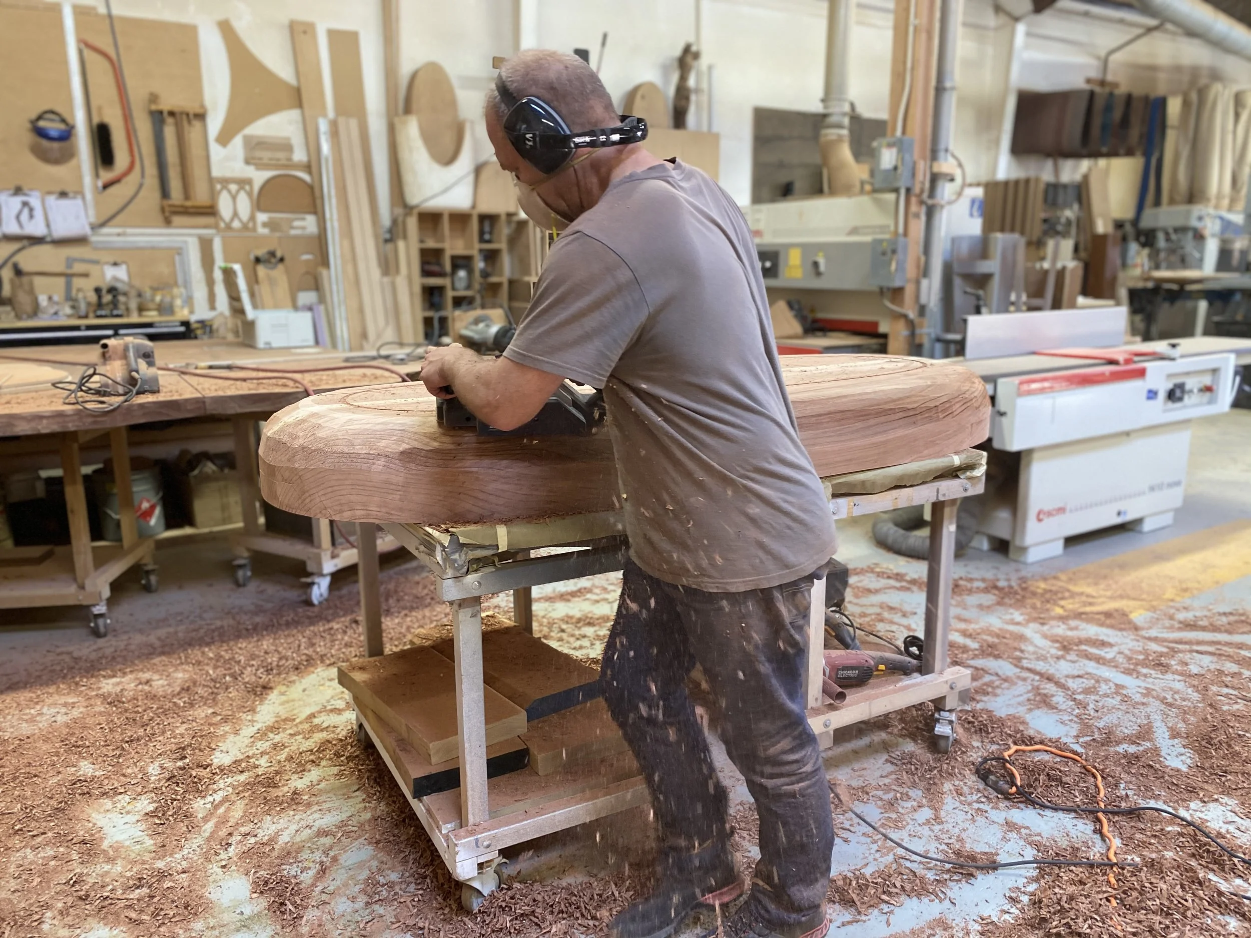 A man working on a large wooden piece in a woodworking shop, using a power tool while wearing protective ear muffs.