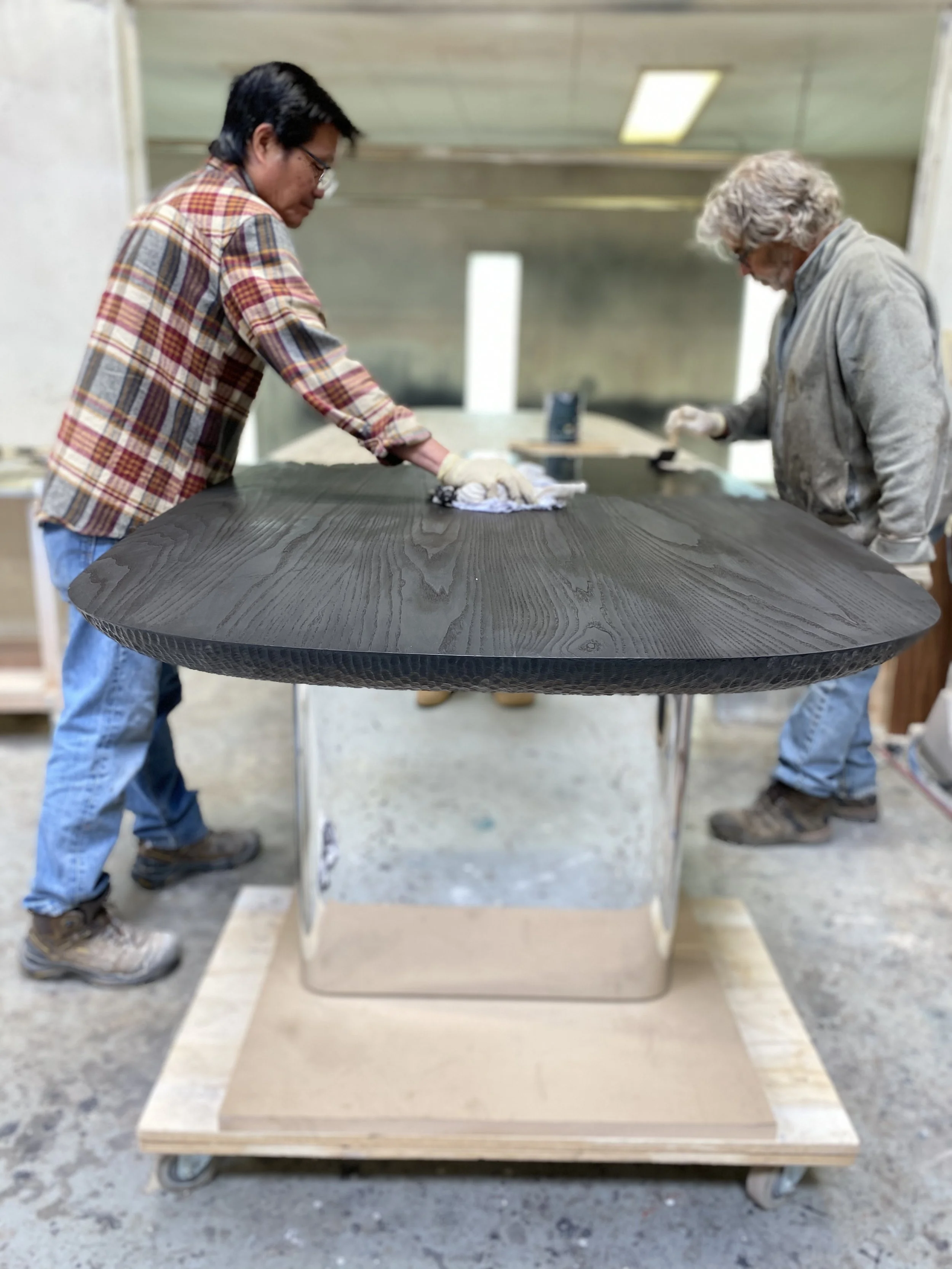 Two people are polishing a large black wooden tabletop supported by a reflective metal base in a workshop.