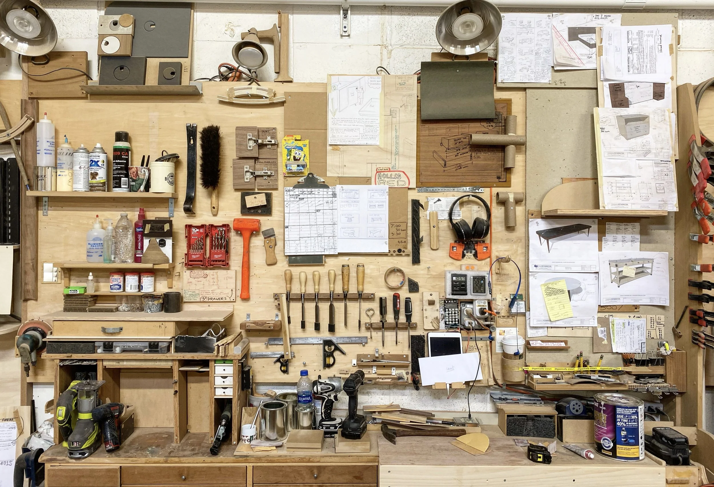 A woodworking workshop with various tools and supplies organized on a wall-mounted pegboard and shelves, including screwdrivers, hammers, clamps, power drills, tape measures, plans, and containers of screws and paints.