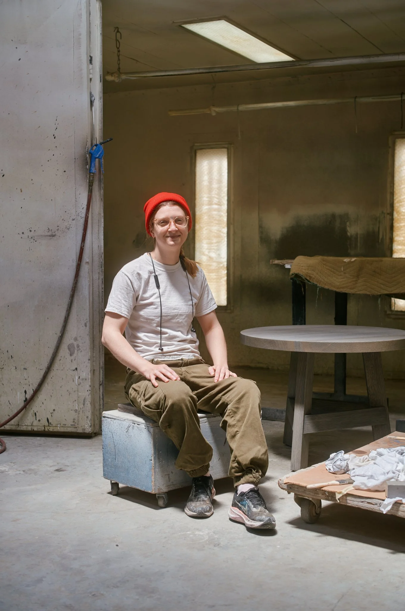 Person sitting on a small metal chair inside a workshop or studio. They are wearing a red beanie, white T-shirt, khaki pants, and sneakers. There are tools and furniture around, with an industrial and workshop-like environment.