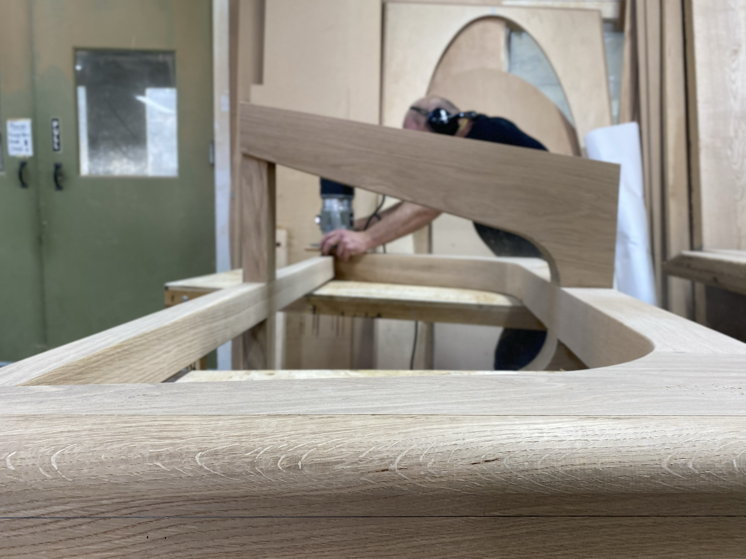 A person wearing safety glasses and a black shirt working on a woodworking project in a workshop, cutting or shaping a piece of wood that is part of a larger wooden structure.