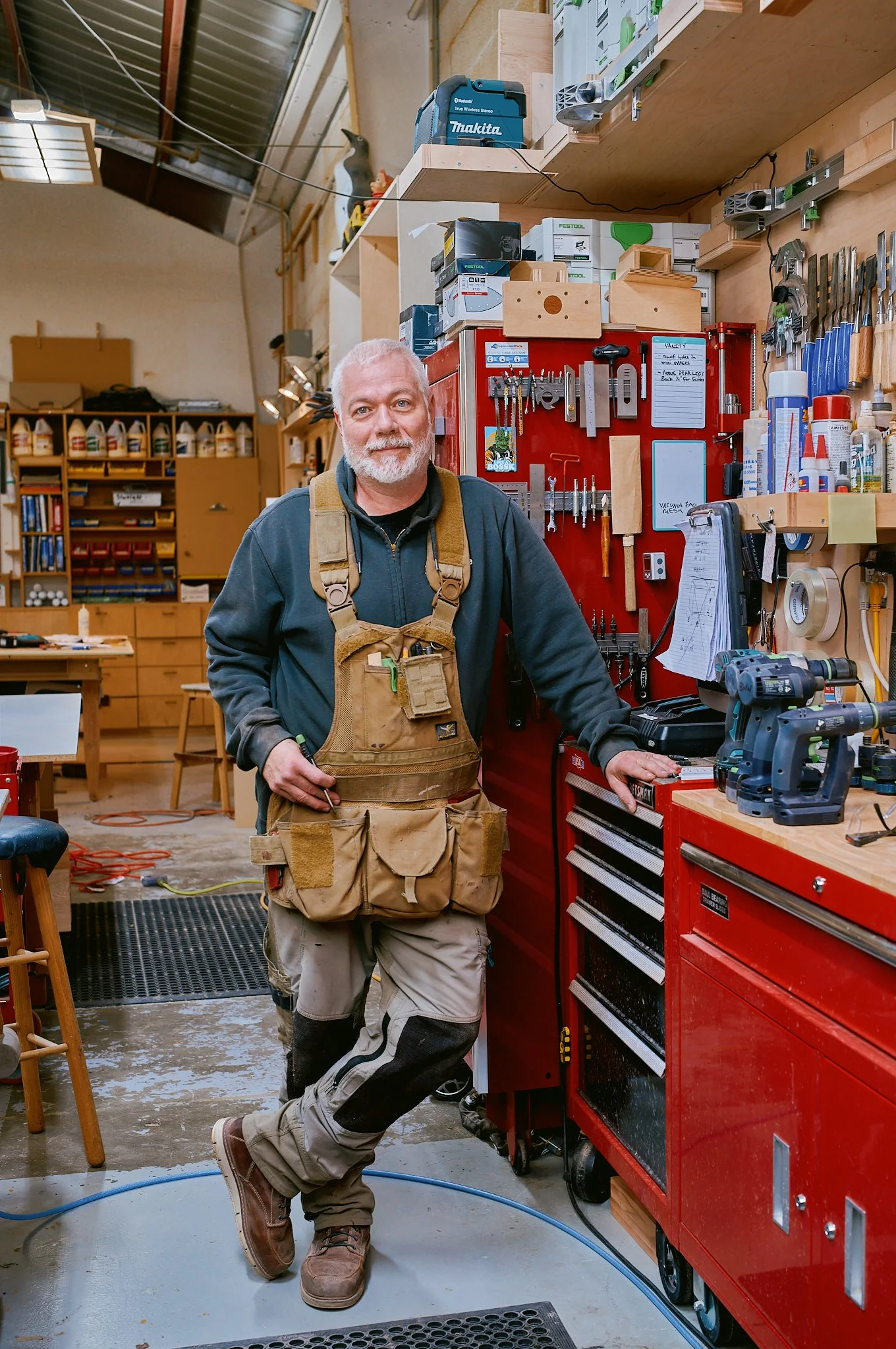 A man in a workshop standing in front of a workbench with tools, wearing work clothing, including a tool belt and overalls, with a smile.