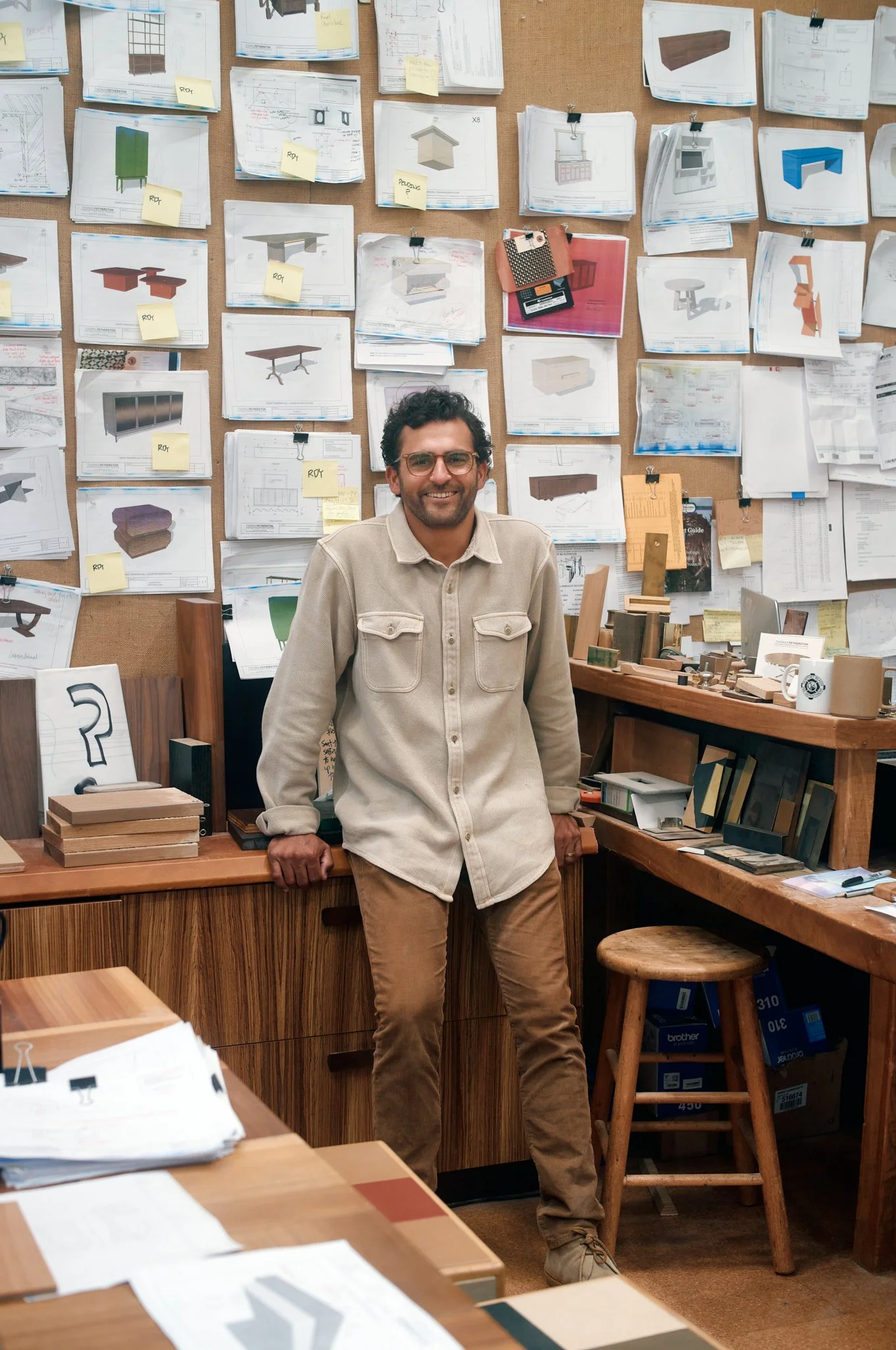 A man with glasses and dark curly hair, smiling, standing in a woodworking or furniture design studio, surrounded by sketches, plans, and samples of furniture and wood materials on a wall and work surfaces.