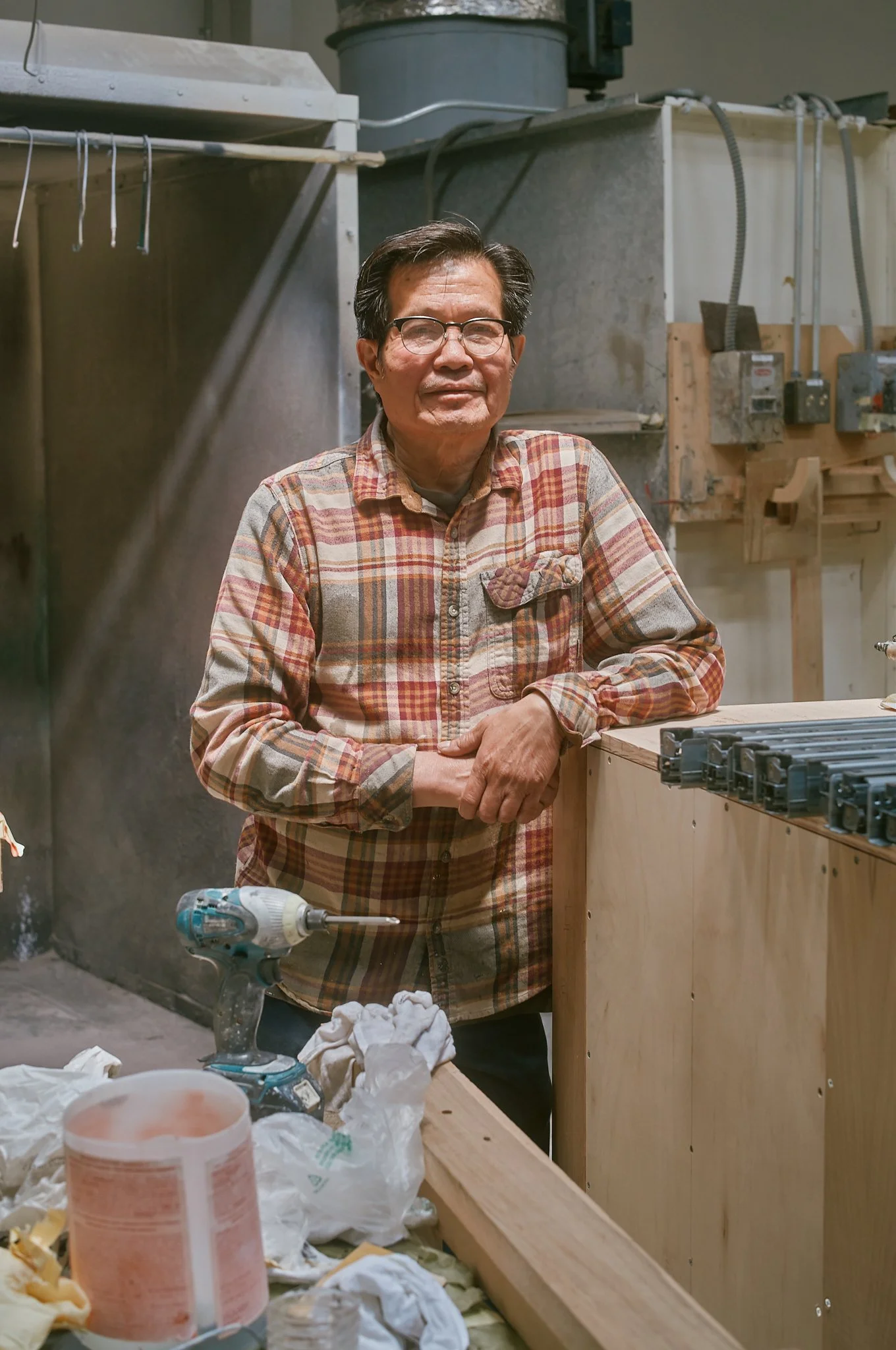 A man in glasses and a plaid shirt standing in a wood workshop with tools and lumber around him.