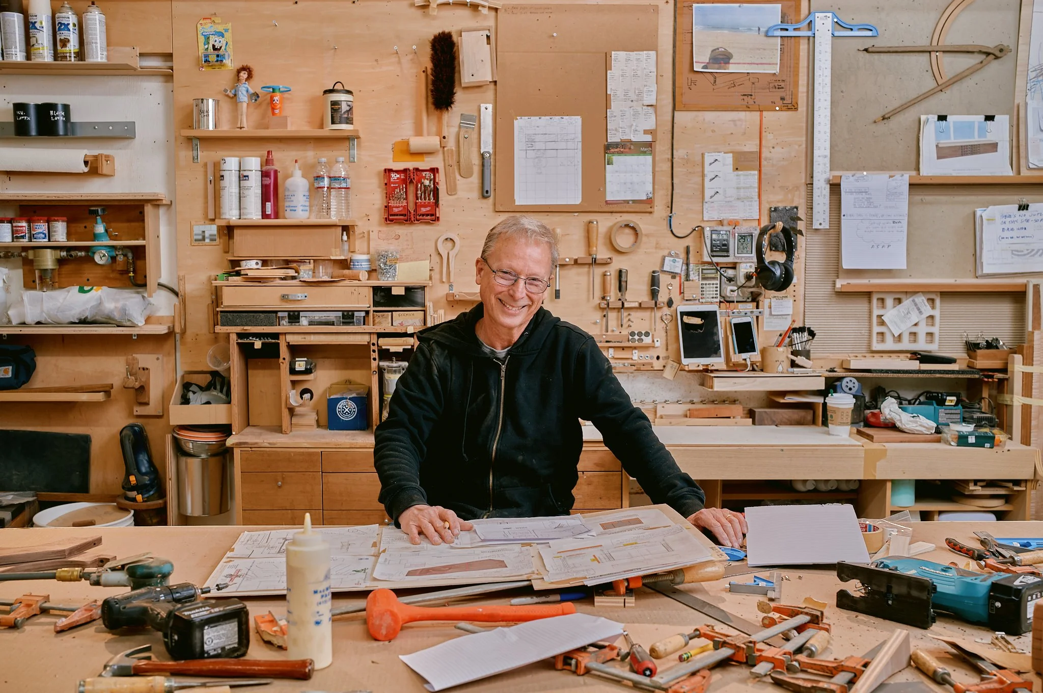A smiling man in a black hoodie working in a woodworking workshop. The workshop has a wooden wall with various tools, supplies, and papers pinned up. The workbench in front of him has plans, tools, and a glue bottle.