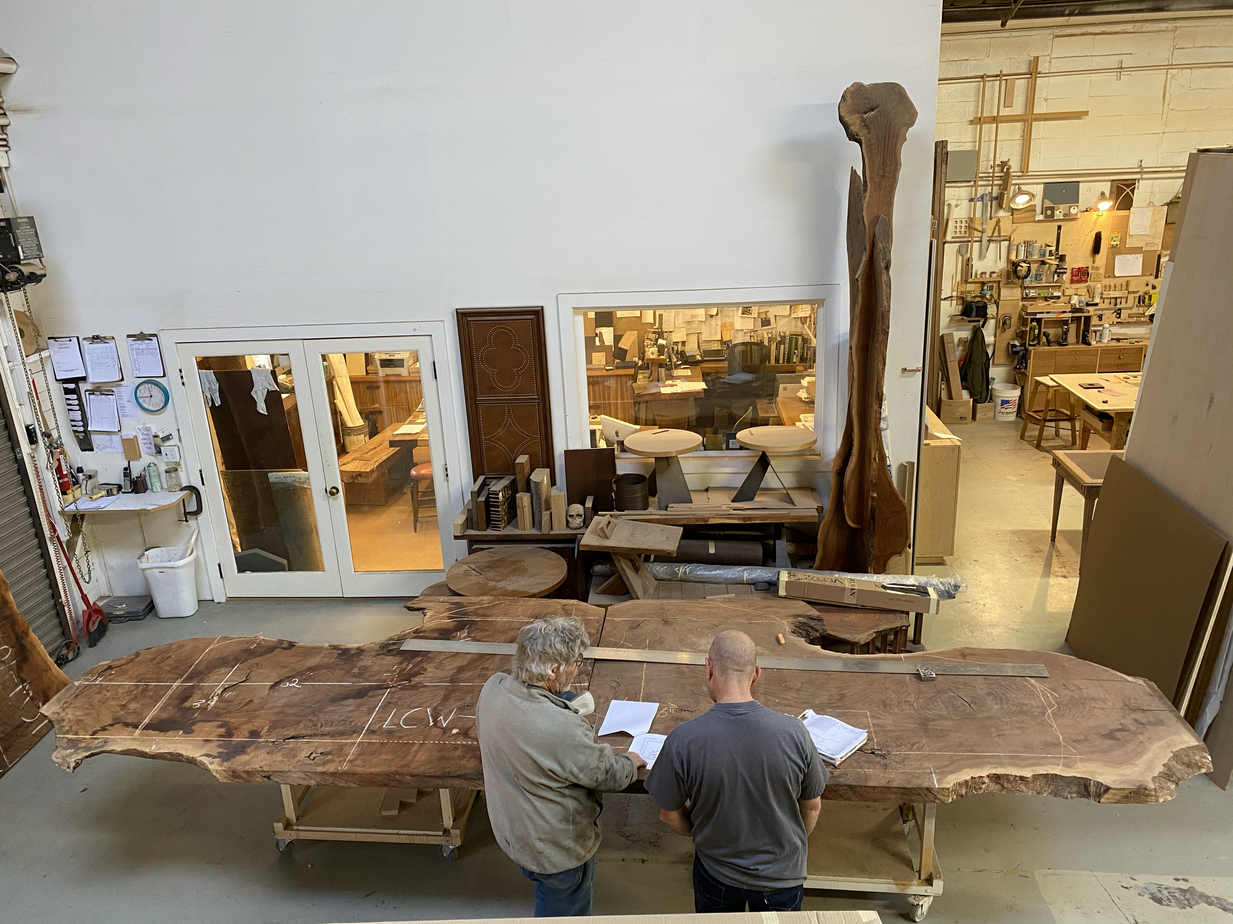 Two men examine a large, live-edge wood slab in a woodworking shop, surrounded by woodworking tools, shelves, and workshop furniture.
