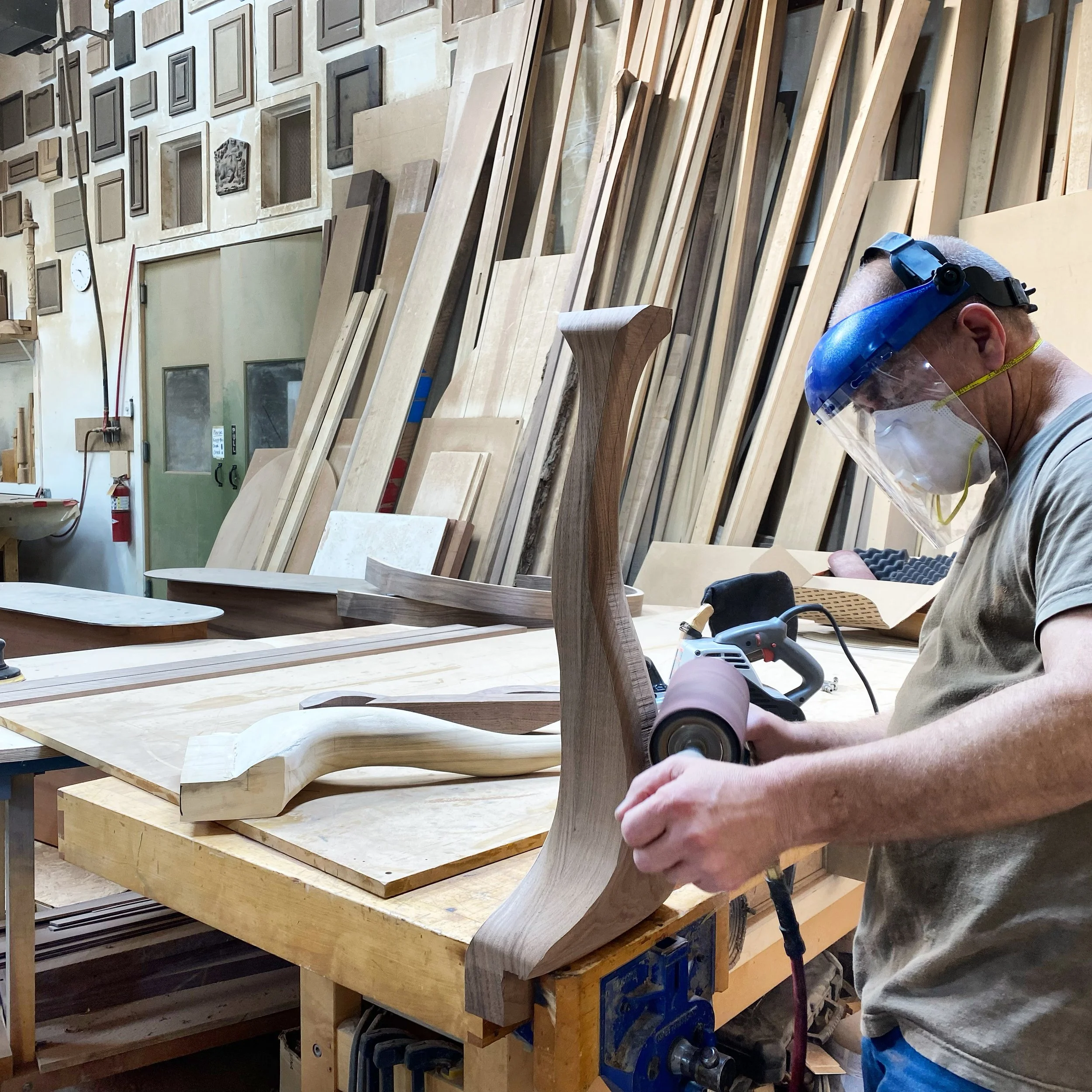 A man wearing a face shield and mask is working on a woodworking project, sanding a curved wooden piece at a workbench in a woodworking shop filled with various wooden planks and furniture pieces.