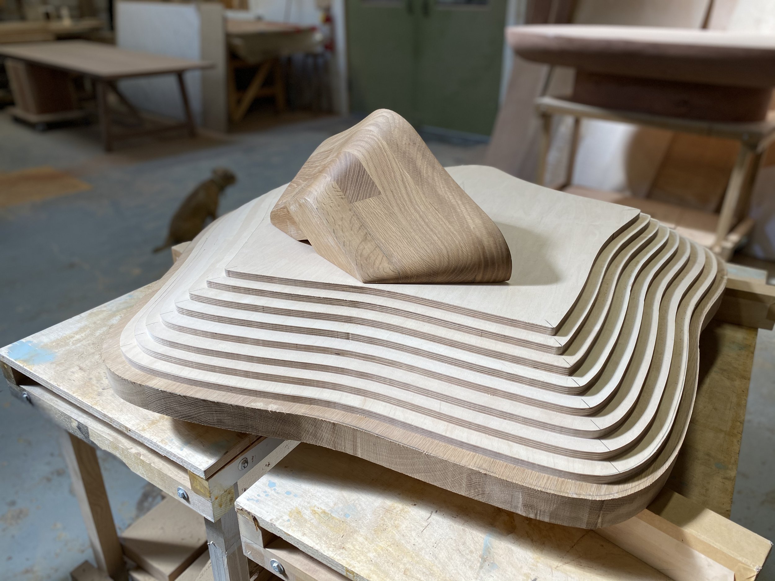 A stack of layered wooden pieces with a carved triangular wooden sculpture on top, situated on a workbench in a woodworking shop.