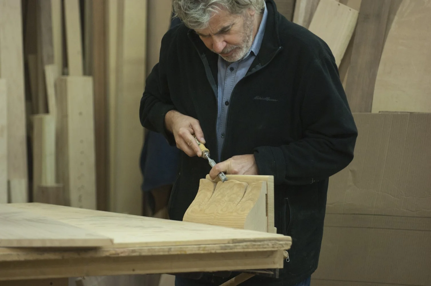 A man with gray hair and beard shaping wood with a chisel in a woodworking shop filled with wooden planks.