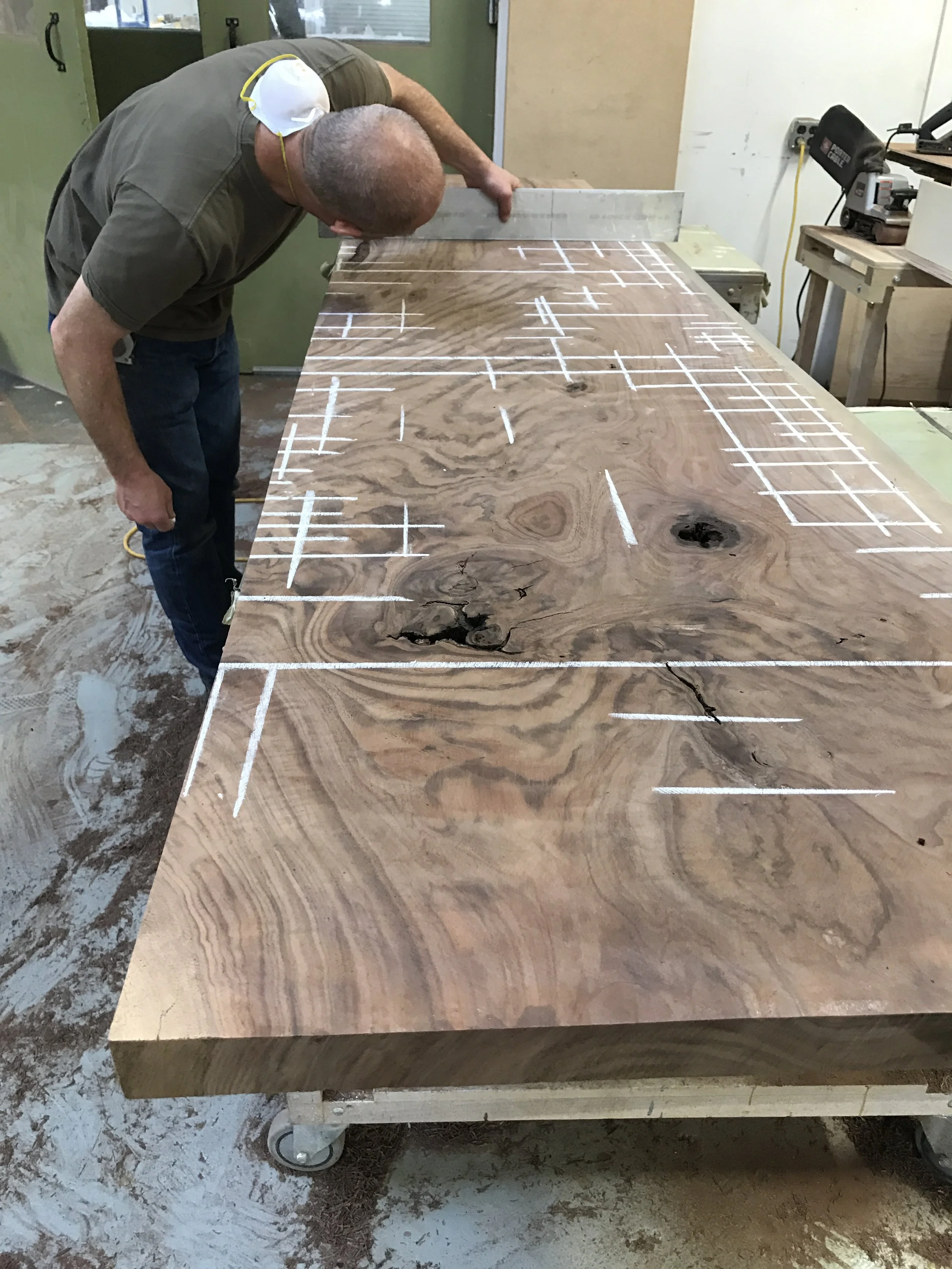 A man inspecting a large wooden workpiece with white markings in a woodworking shop.