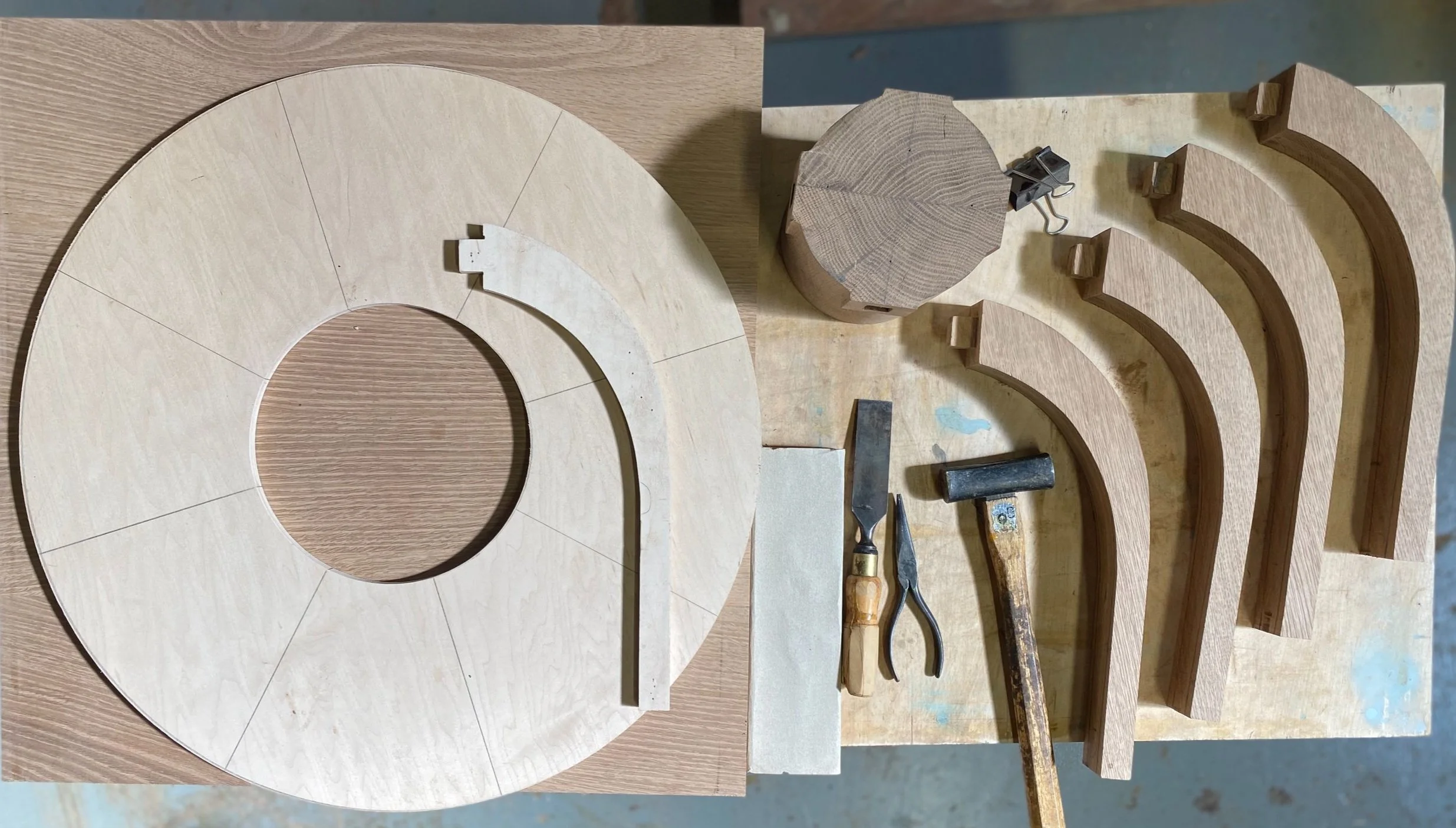 Woodworking tools and pieces of wood on a workbench, including a large circular piece, curved wooden parts, a mallet, chisels, and clamps.