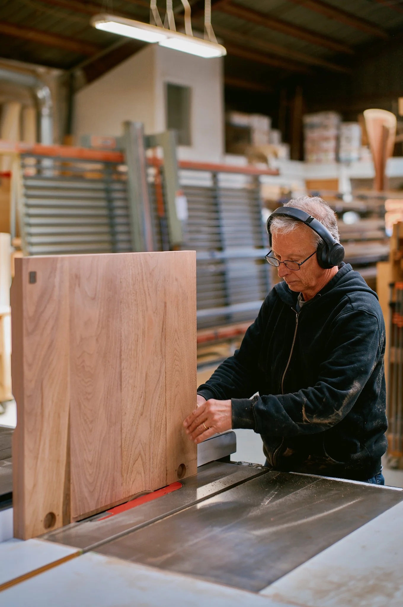 An older man wearing glasses, a black hoodie, and headphones uses a table saw to cut a piece of wood in a woodworking shop.