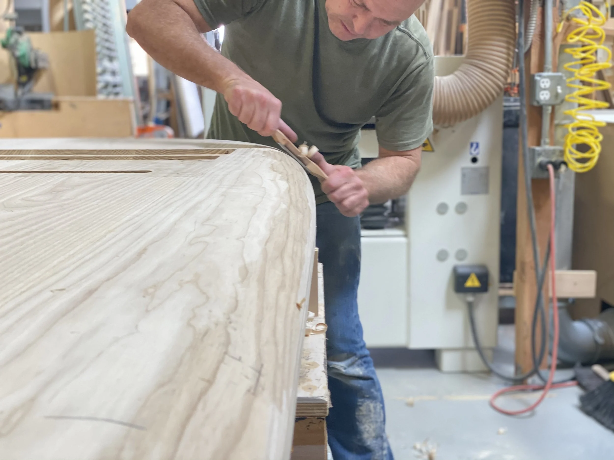 A man is woodworking, carving or sanding a piece of light-colored wood with a chisel or carving tool in a workshop.