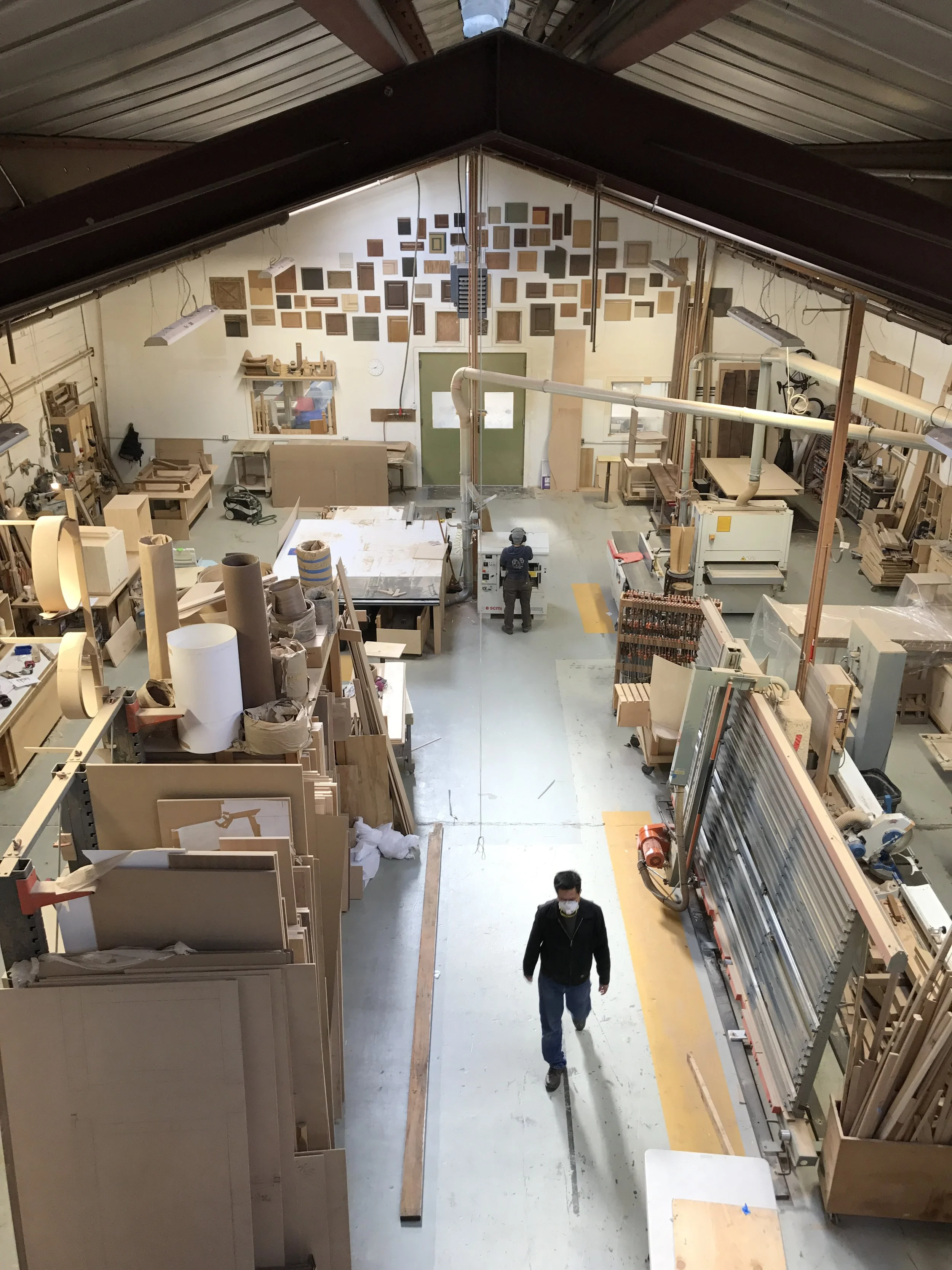 Woodworking workshop with two people wearing masks, one walking towards the camera and another operating machinery. The workshop is filled with wooden boards, tools, and equipment, with a decorative wall installation of various wood samples on the ba