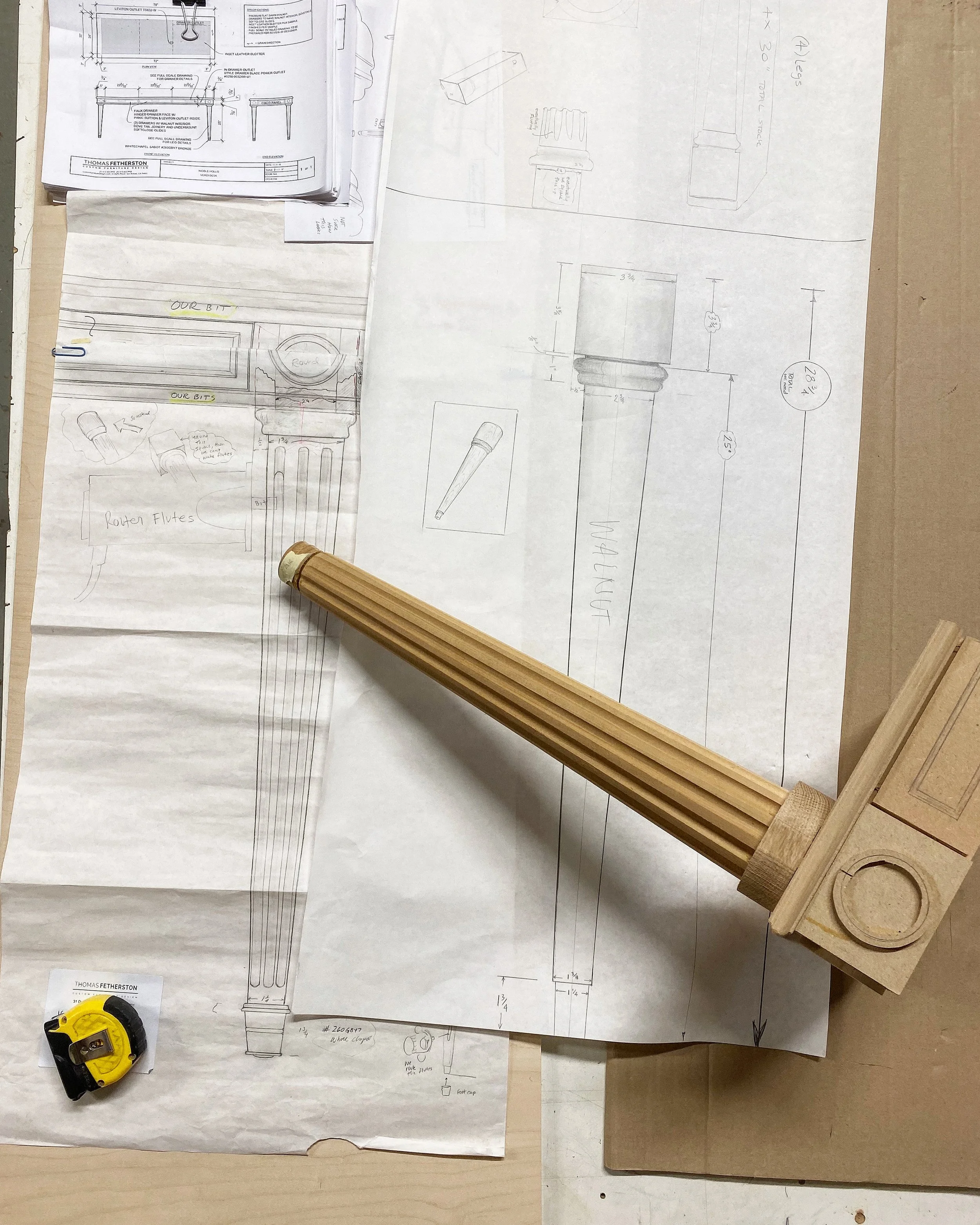 Blueprints and sketches for a furniture piece, possibly a table or lamp, with detailed measurements and design notes. There are 3D model sketches, handwritten notes, and a wooden component with slats attached to a block, resting on the blueprints.
