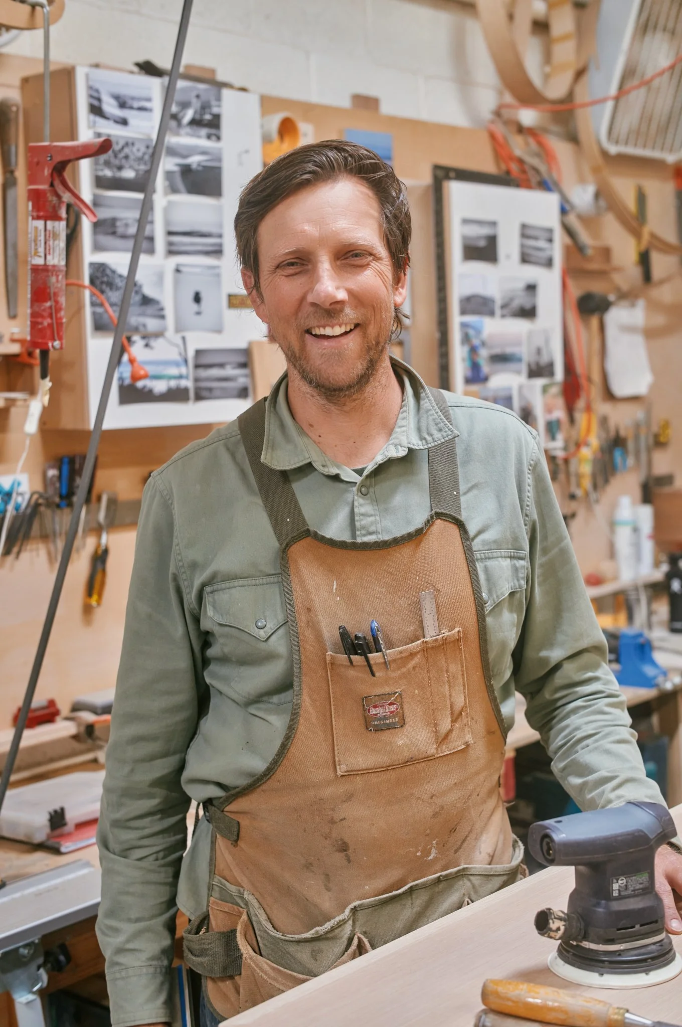 A smiling man standing in a woodworking shop, wearing a green shirt and a brown apron with pens and a ruler in the front pocket. Tools and photos are visible on the wall behind him, with woodworking equipment on the table and in the background.