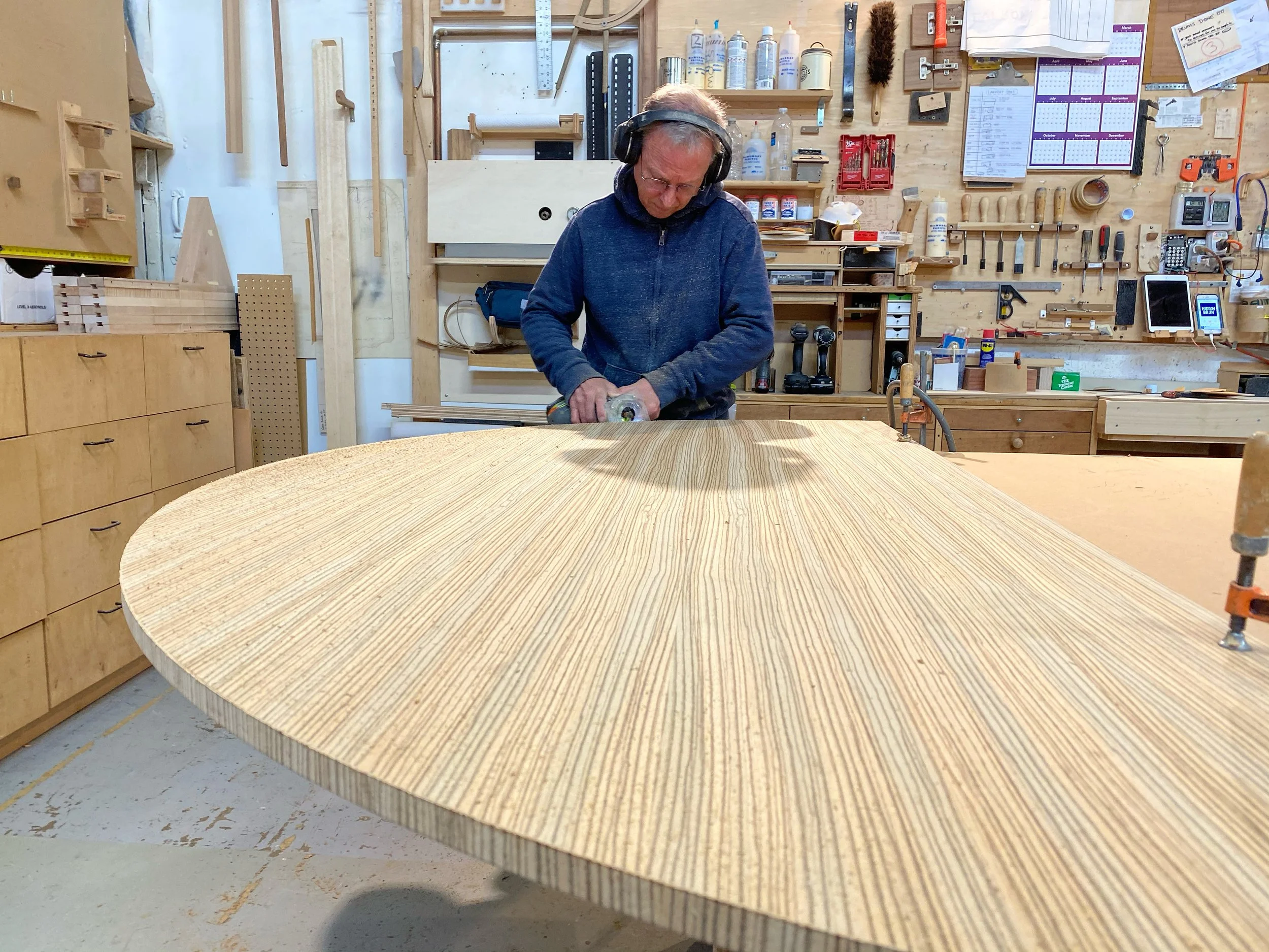 A man wearing safety headphones and a blue jacket is using a power sander on a large, curved piece of wood in a woodworking shop.
