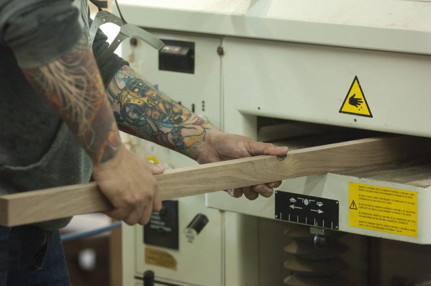 A person with colorful tattoos on their arms working with a woodworking machine, guiding a piece of wood through it.