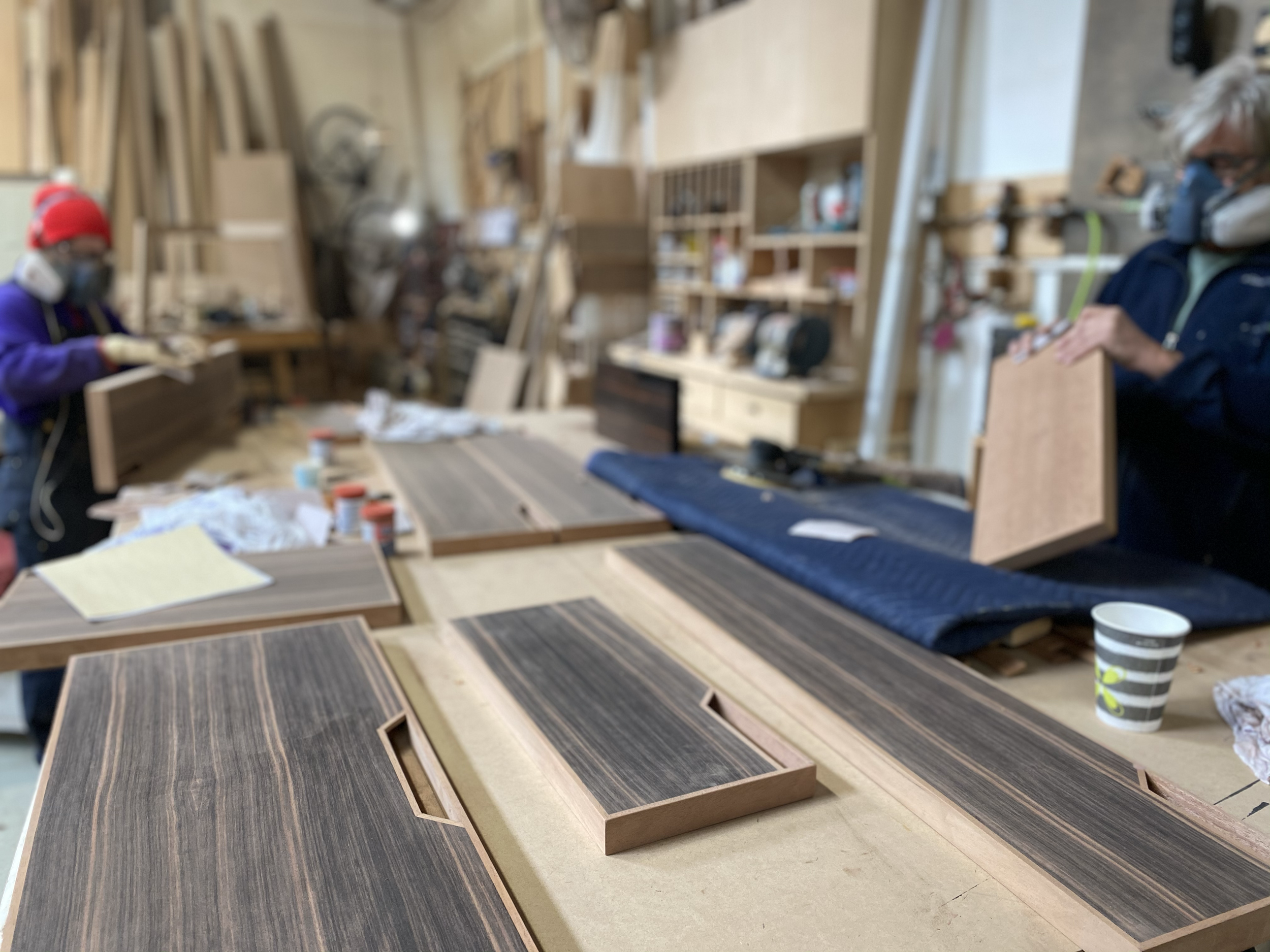 Two people working on woodworking projects in a workshop with wooden panels, tools, and materials on a large table.