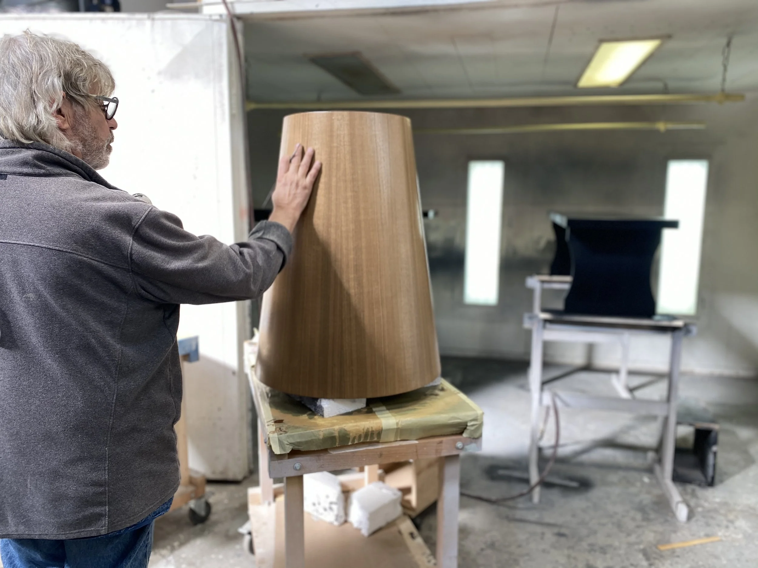 A man with gray hair, glasses, and a gray jacket touching a large, conical wooden object on a work table inside a workshop or factory.