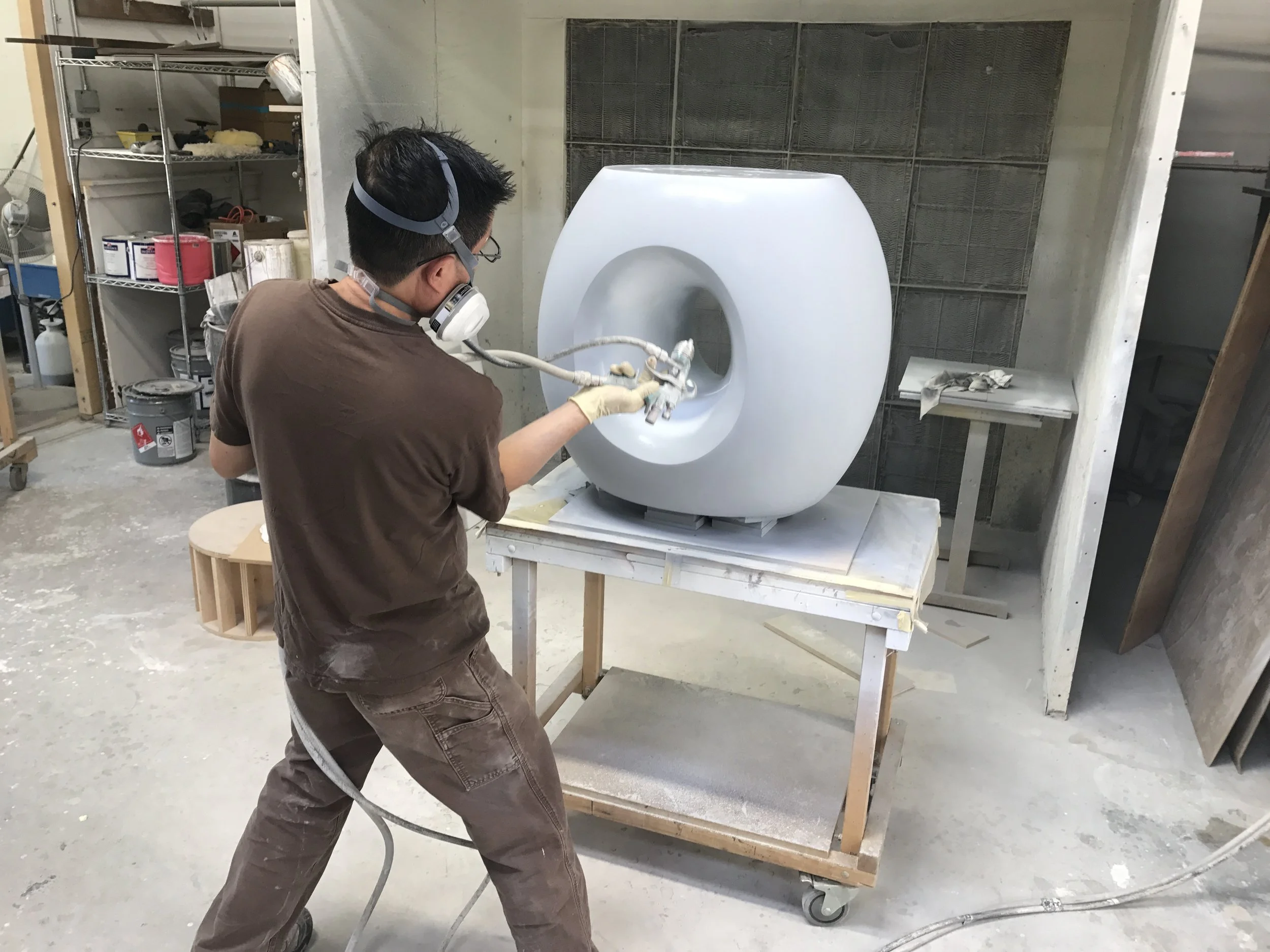 Person wearing a mask and gloves spray painting or finishing a large, white, modern-looking ceramic or stone toilet on a workbench in a workshop.