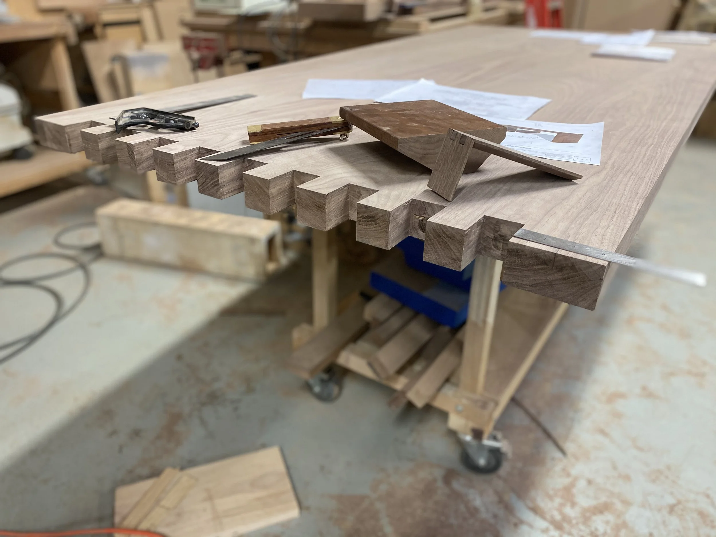 Woodworking workshop with a large worktable, woodworking tools like clamps and chisels, and pieces of wood, including a large finished piece with dovetail joints on a mobile cart.