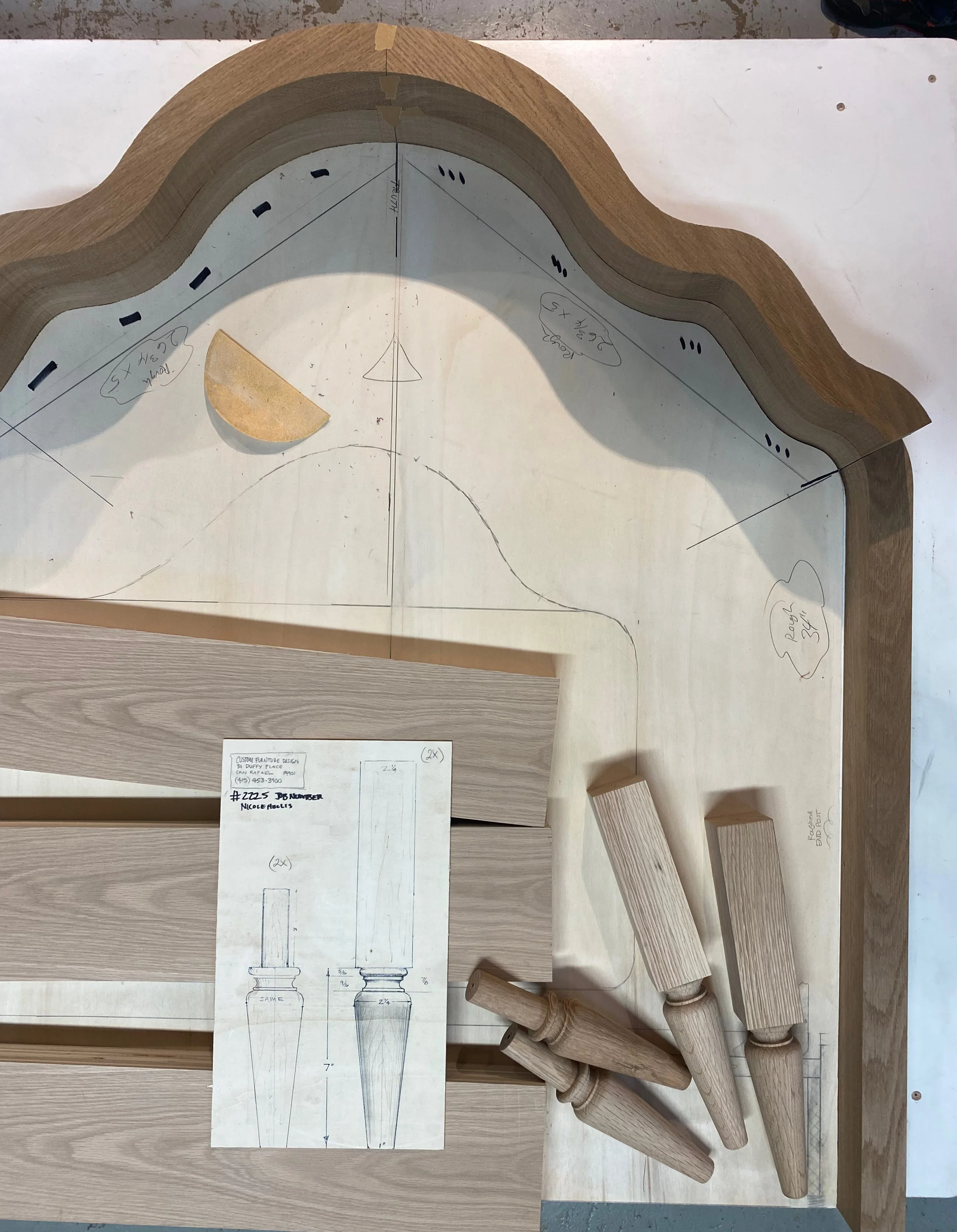 Wooden furniture design with a curved headboard pattern sketched on a light-colored surface. Several wooden bed posts with turned design are placed on the right side of the image. A paper plan with measurements and sketches of bedposts is positioned 