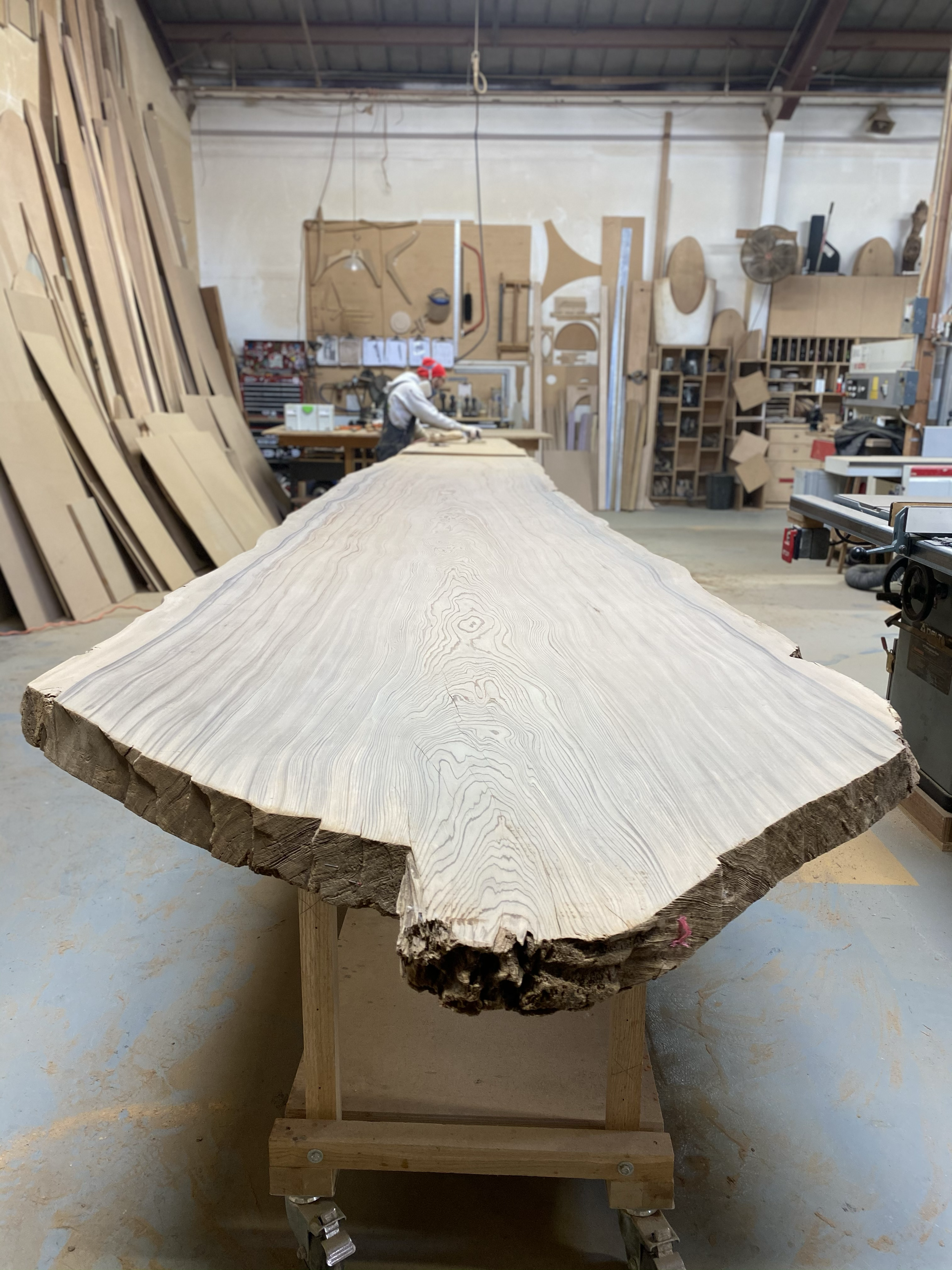 A large, natural wood slab with visible growth rings and natural edges, placed on a wheeled wooden stand in a woodworking shop. In the background, a person wearing a red hat and white hoodie is working at a workbench surrounded by various woodworking