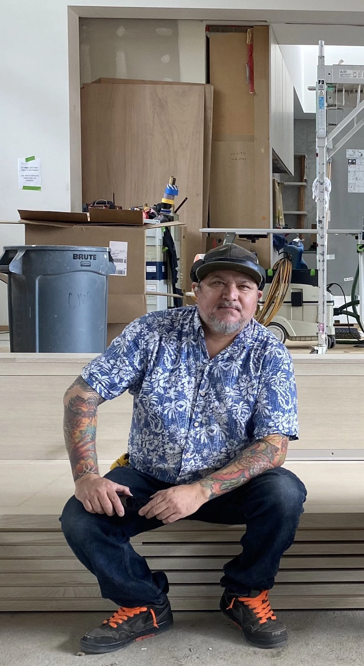 A man with tattoos on his arms sitting on a wooden surface in a workshop, wearing a patterned blue Hawaiian shirt, black pants, black and orange shoes, a black baseball cap, and earplugs, with construction materials and tools in the background.