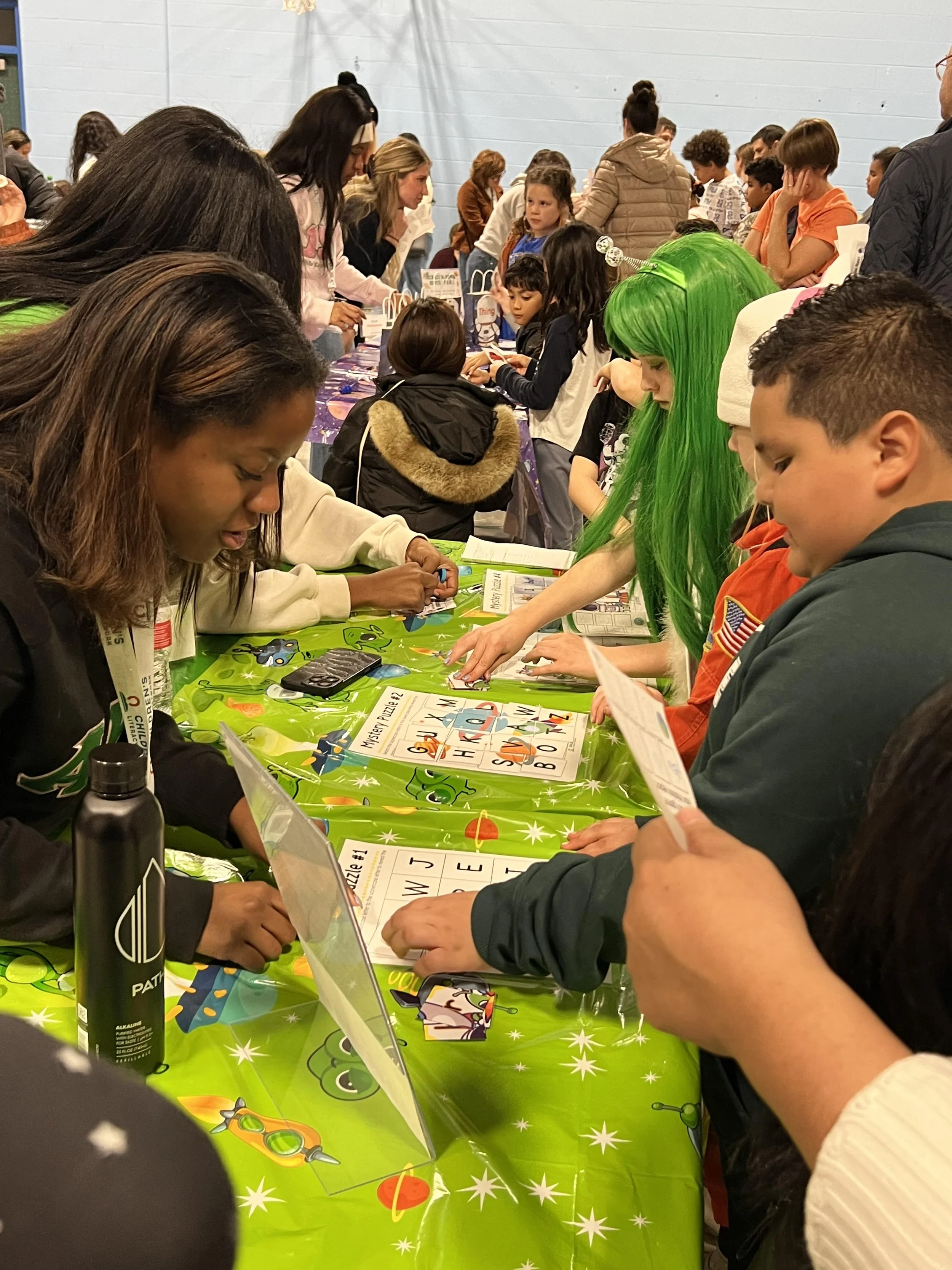 Children playing tabletop literacy games in a library with adult volunteers supervising.