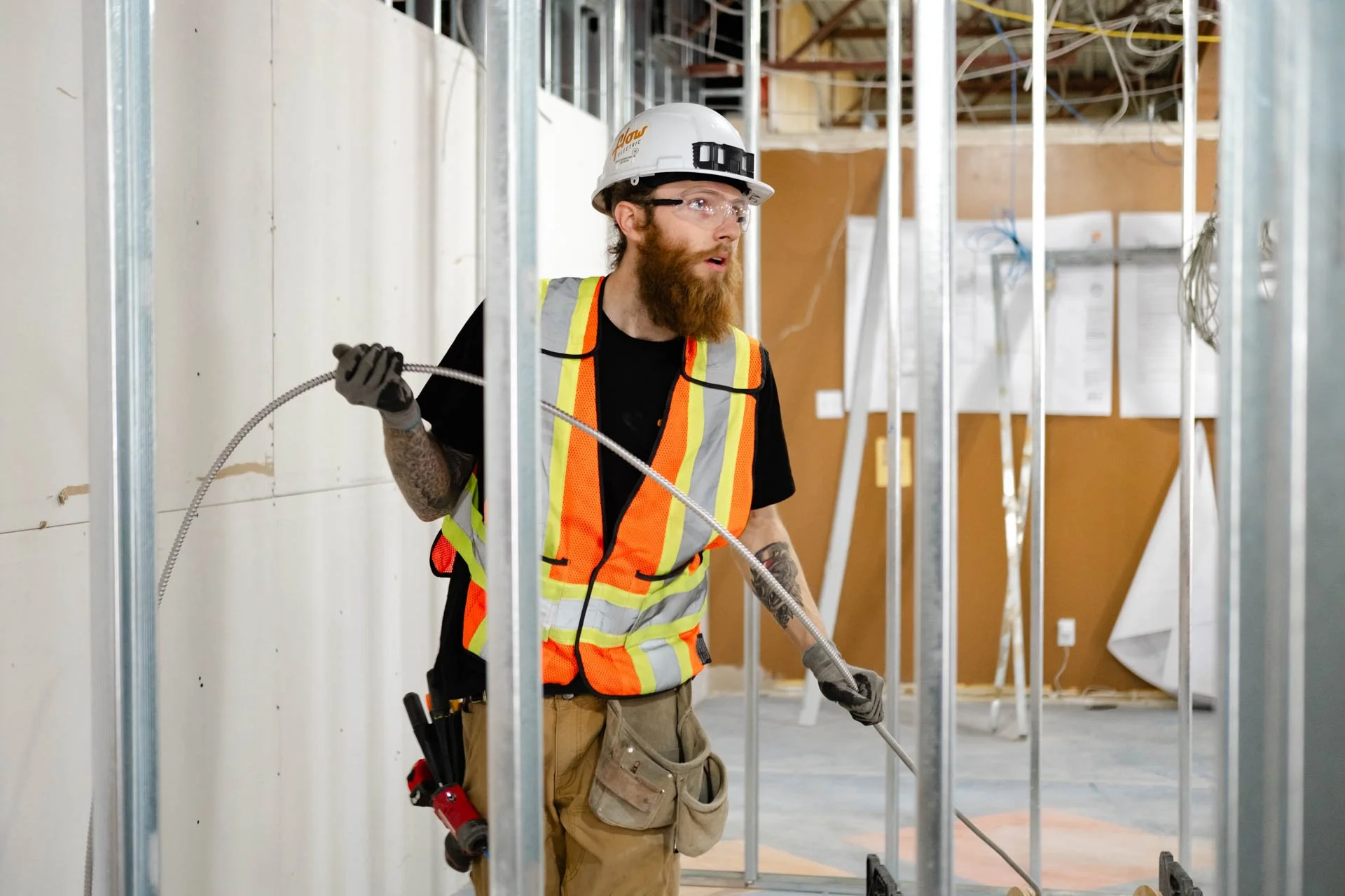 Construction worker with a beard and tattoos, wearing a white safety helmet, glasses, a high-visibility safety vest, gloves, and carrying tools, is working inside a building under construction, framing metal studs are visible around him.