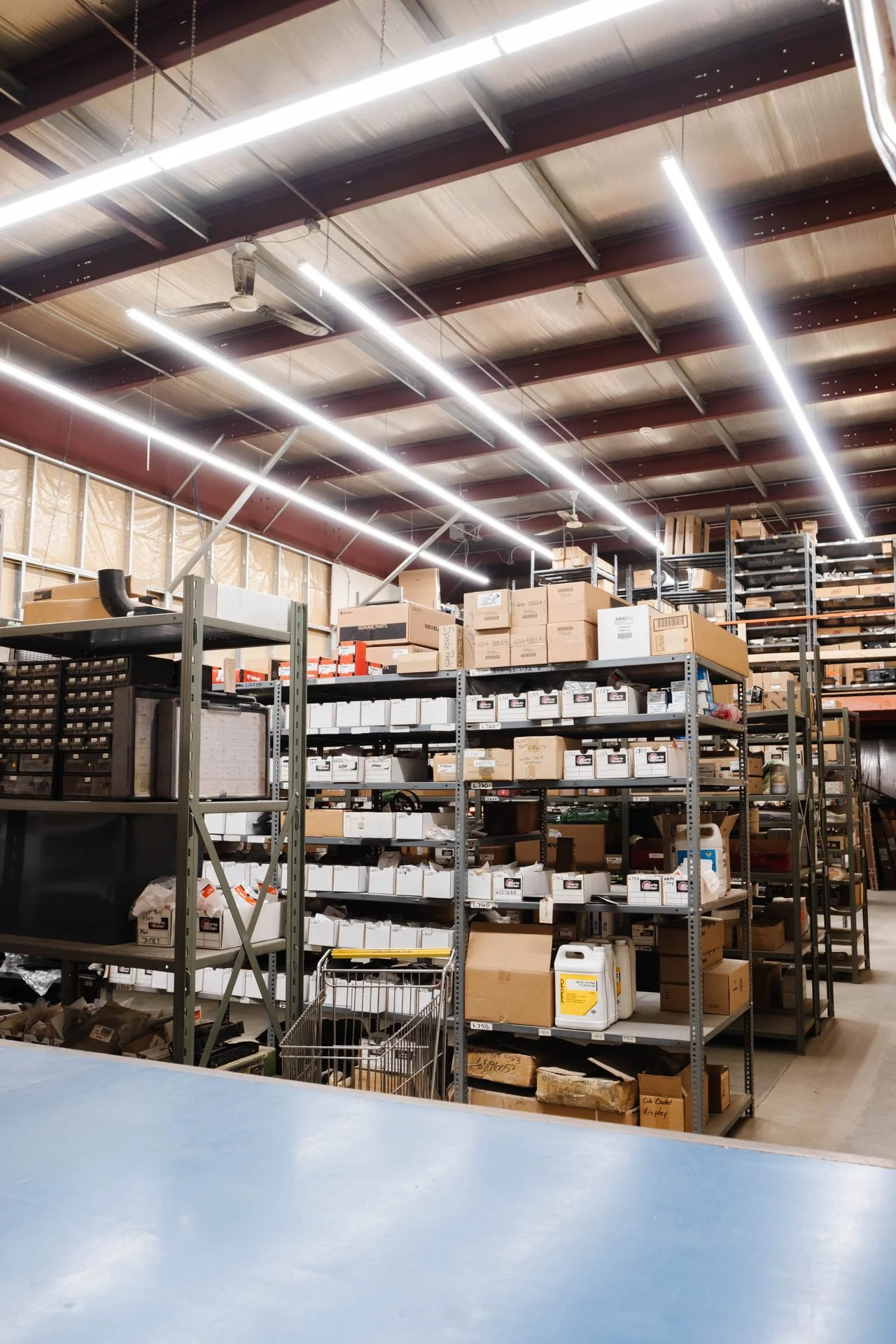 Industrial warehouse with metal shelving units filled with boxes and containers, fluorescent ceiling lights, and a ceiling fan.