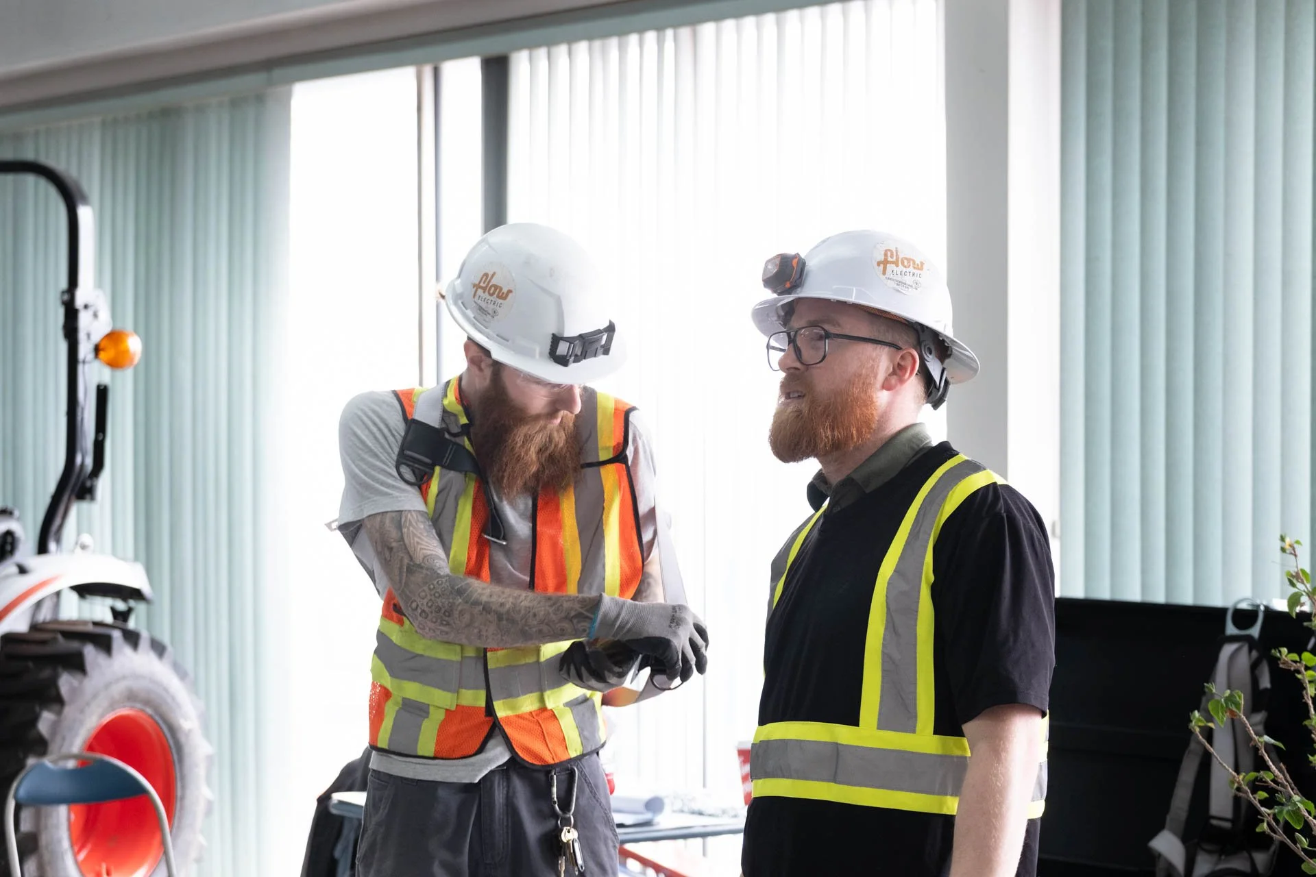 Two male construction workers with safety helmets and reflective vests are having a conversation indoors. One is holding a device, and the other is listening. The background shows windows and some equipment.
